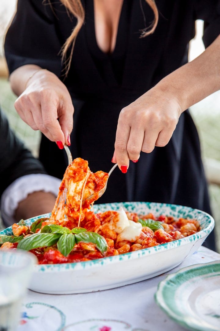 A person serves gnocchi with tomato sauce and basil from a decorative dish onto a plate at a table.