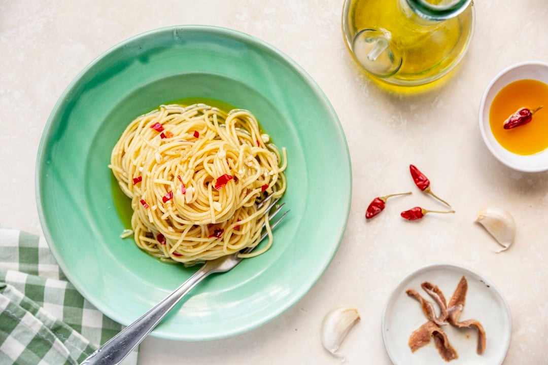 A plate of spaghetti aglio e olio with red chili flakes on a green plate. Nearby are a bottle of olive oil, garlic cloves, a bowl of oil with chilies, and anchovies on a small plate.