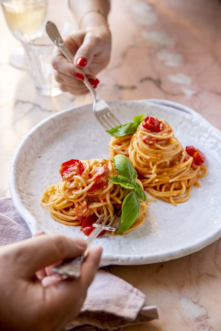 Two people use forks to eat spaghetti with tomato sauce and basil on a white plate, set on a marble table with a glass of water in the background.