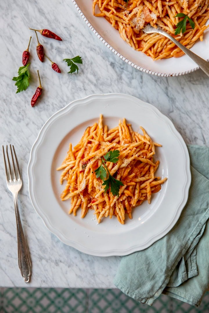A plate of pasta with tomato sauce and a parsley garnish on a marble surface, accompanied by a fork, chili peppers, and a cloth napkin.