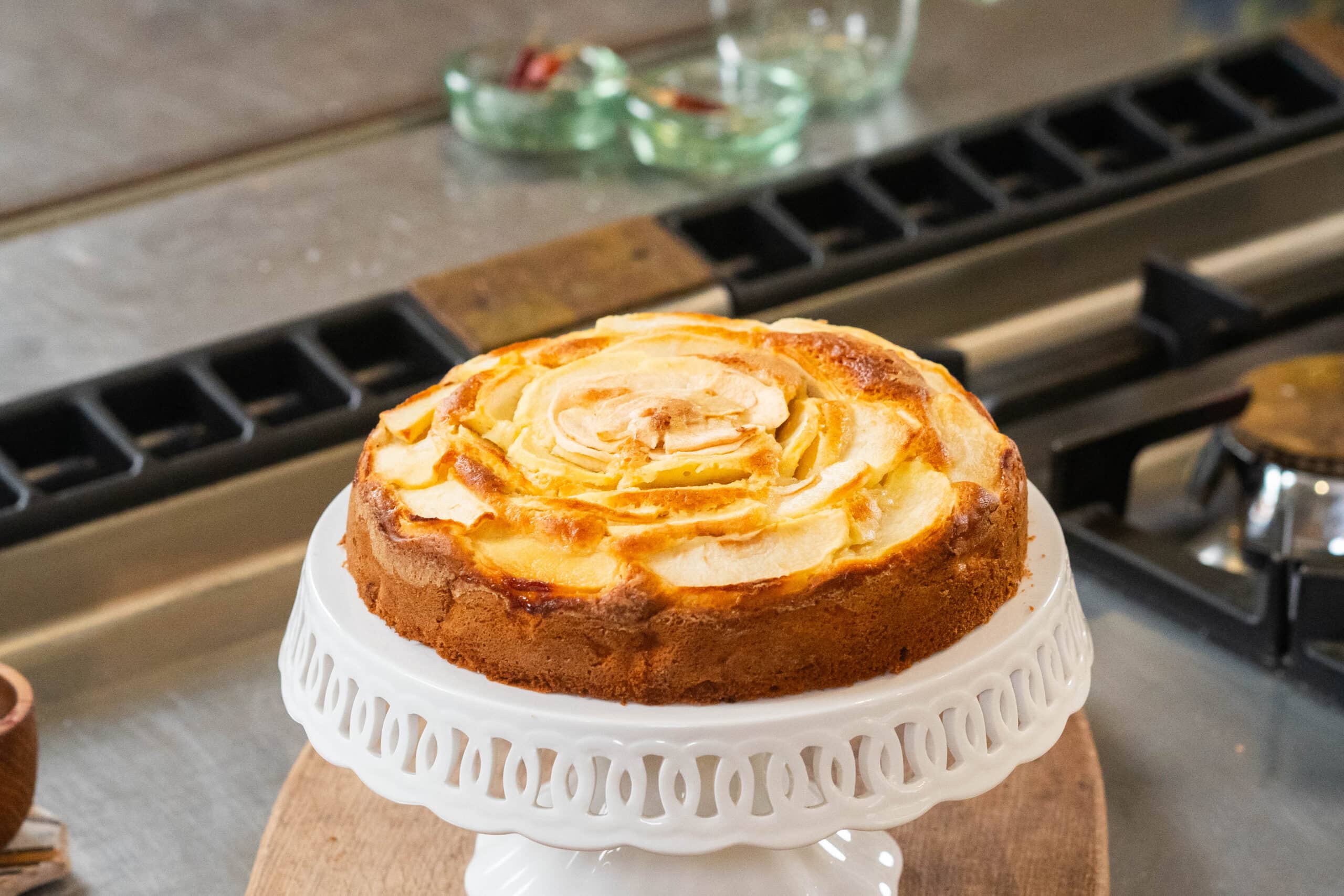 Apple tart on a white cake stand in a kitchen setting with a stove in the background.
