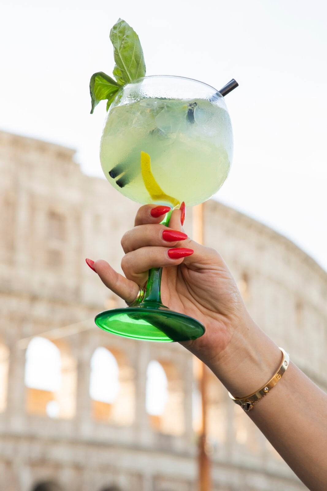 A hand with red nails holds a green-stemmed glass with a light green cocktail, garnished with a lemon twist and basil, in front of a blurred ancient amphitheater background.