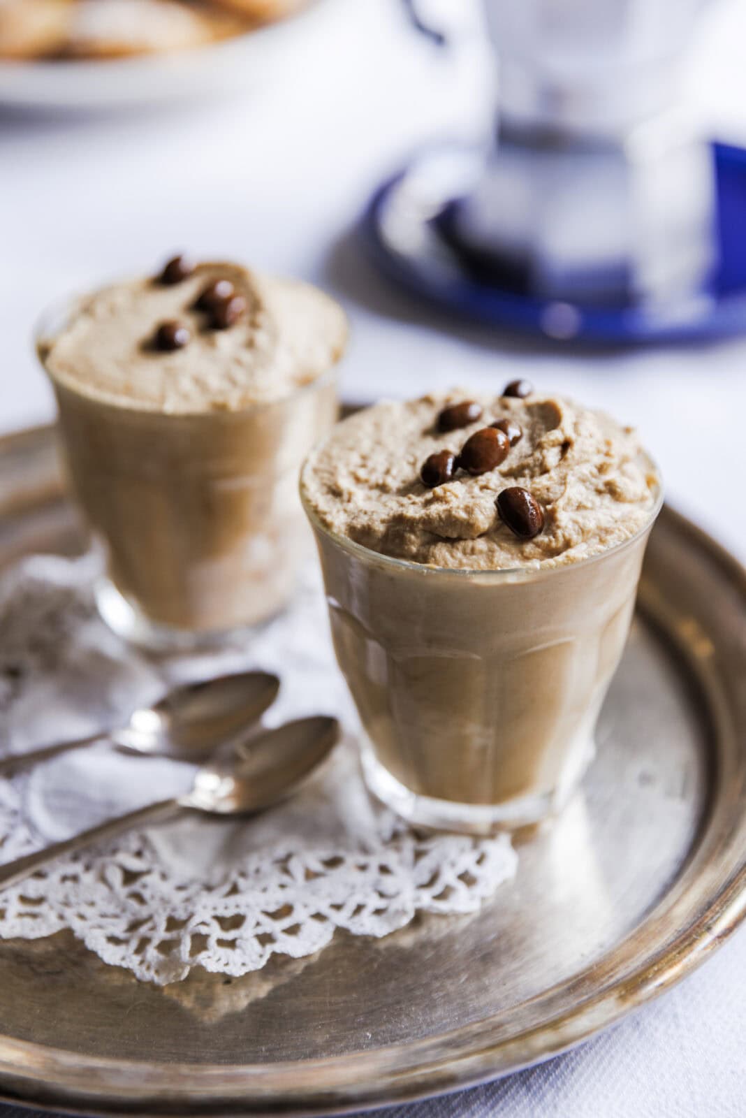 Two glasses of whipped coffee topped with coffee beans on a silver tray, accompanied by two spoons on a lace doily.
