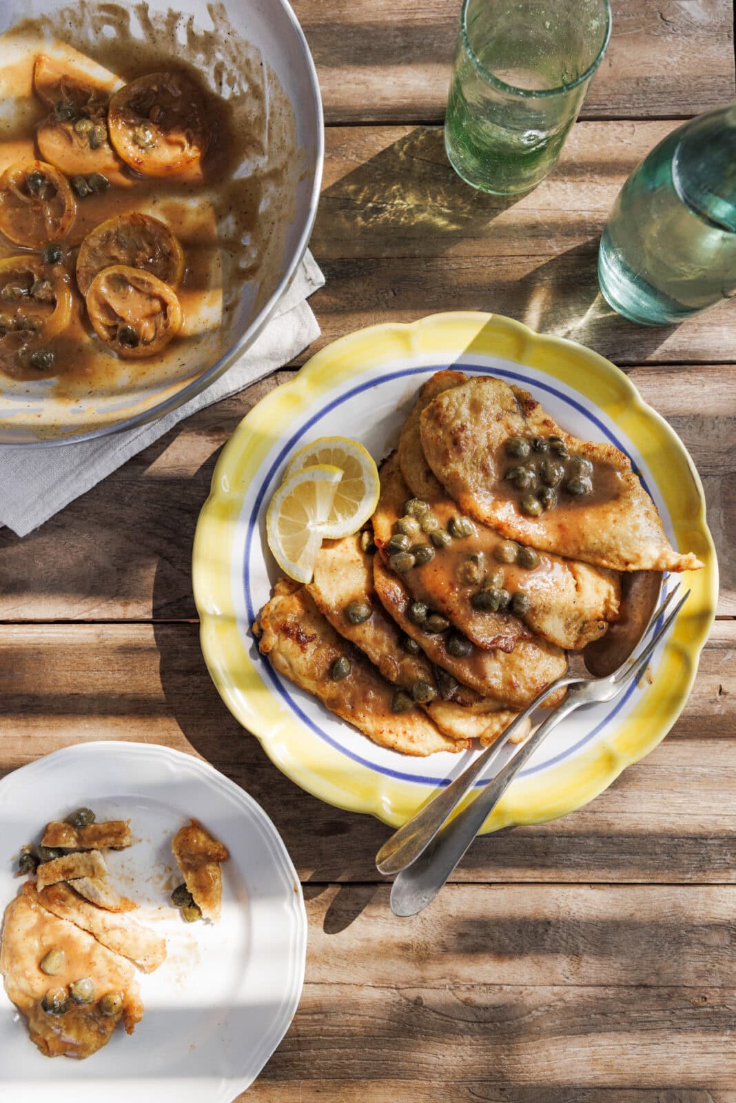Chicken piccata with capers on a yellow-rimmed plate, accompanied by a lemon slice. Nearby are a serving dish, a smaller plate with a piece, and two green glass bottles on a wooden table.