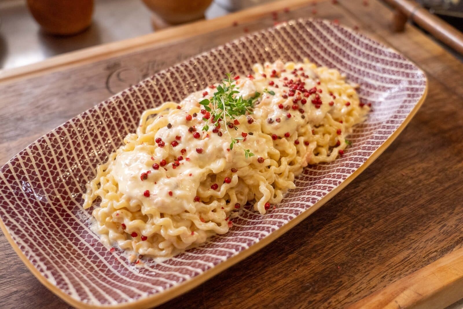 A rectangular plate with pasta topped with creamy sauce, pink peppercorns, and garnished with fresh greens, placed on a wooden surface.