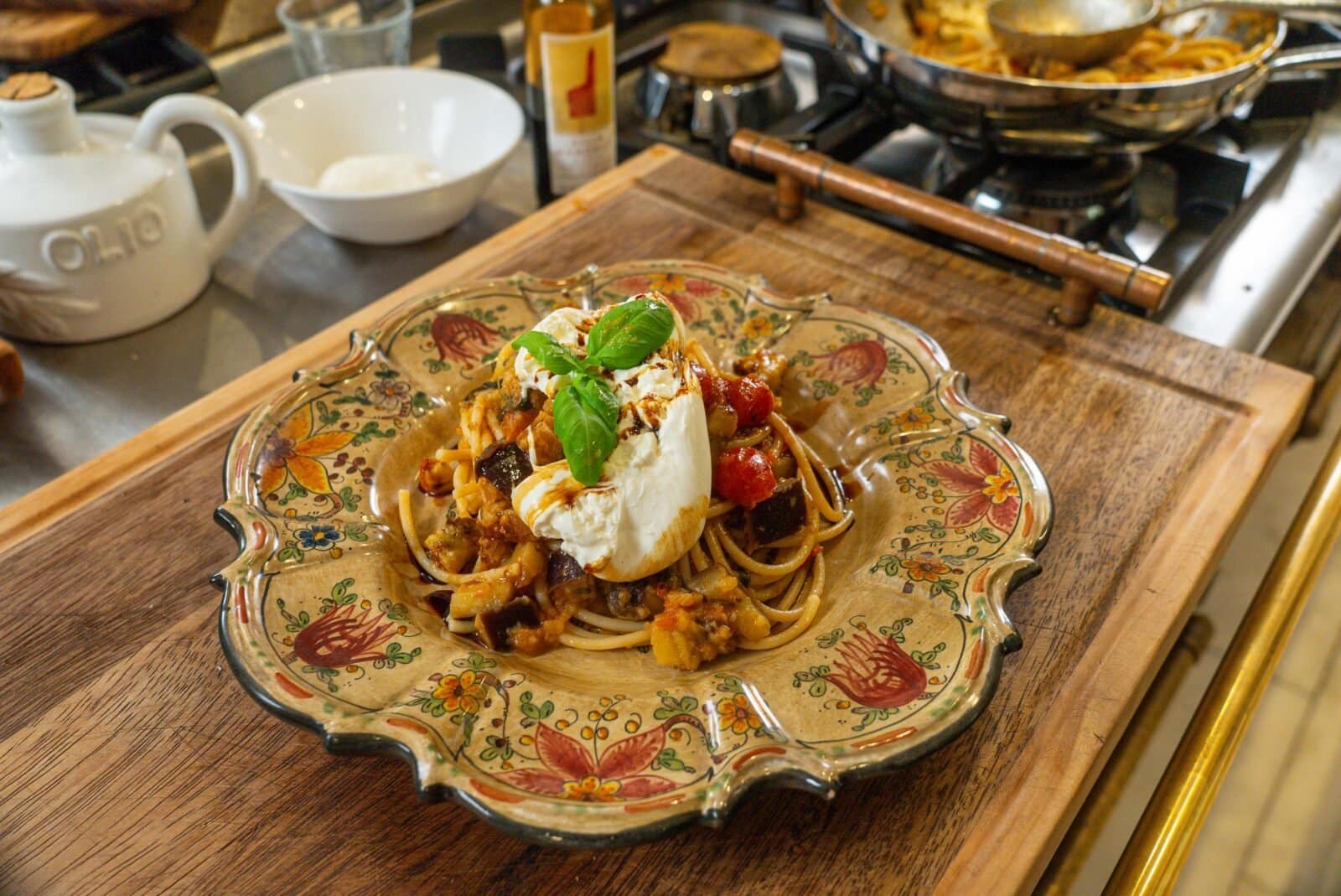 A plate of pasta topped with a ball of burrata, roasted vegetables, and a basil leaf, served on a floral-patterned dish on a wooden cutting board.
