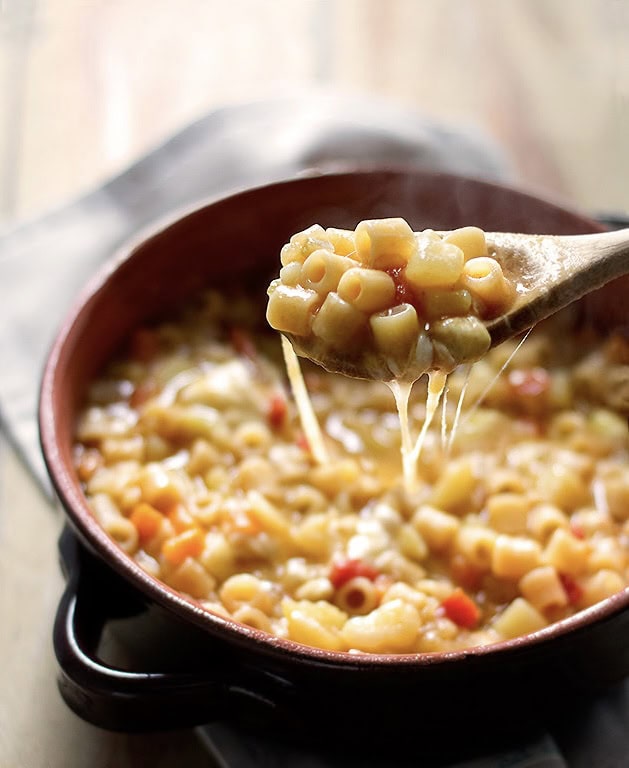 A wooden spoon lifting cheesy pasta from a brown bowl filled with small pasta pieces, tomato bits, and melted cheese.