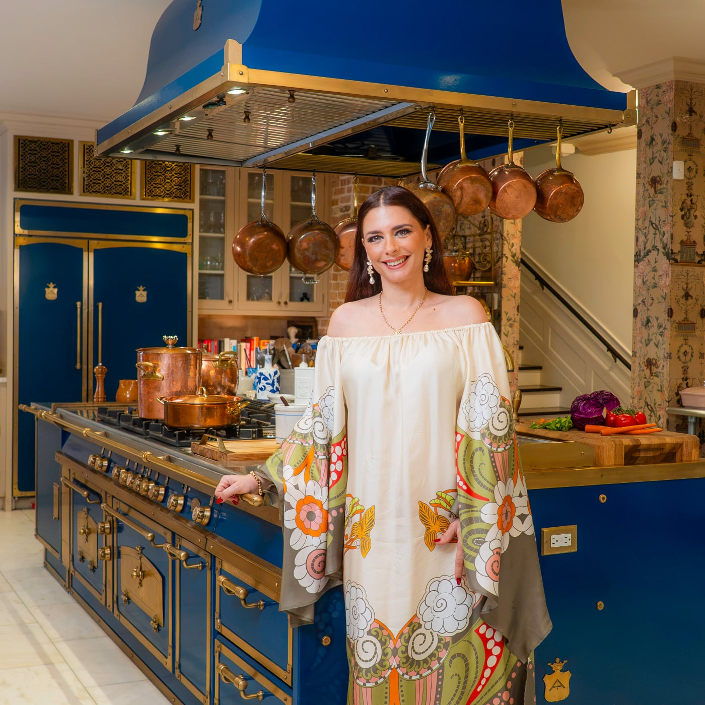 A woman in a cream-colored dress with a floral pattern stands in a blue kitchen with a large range and copper pots hanging above.