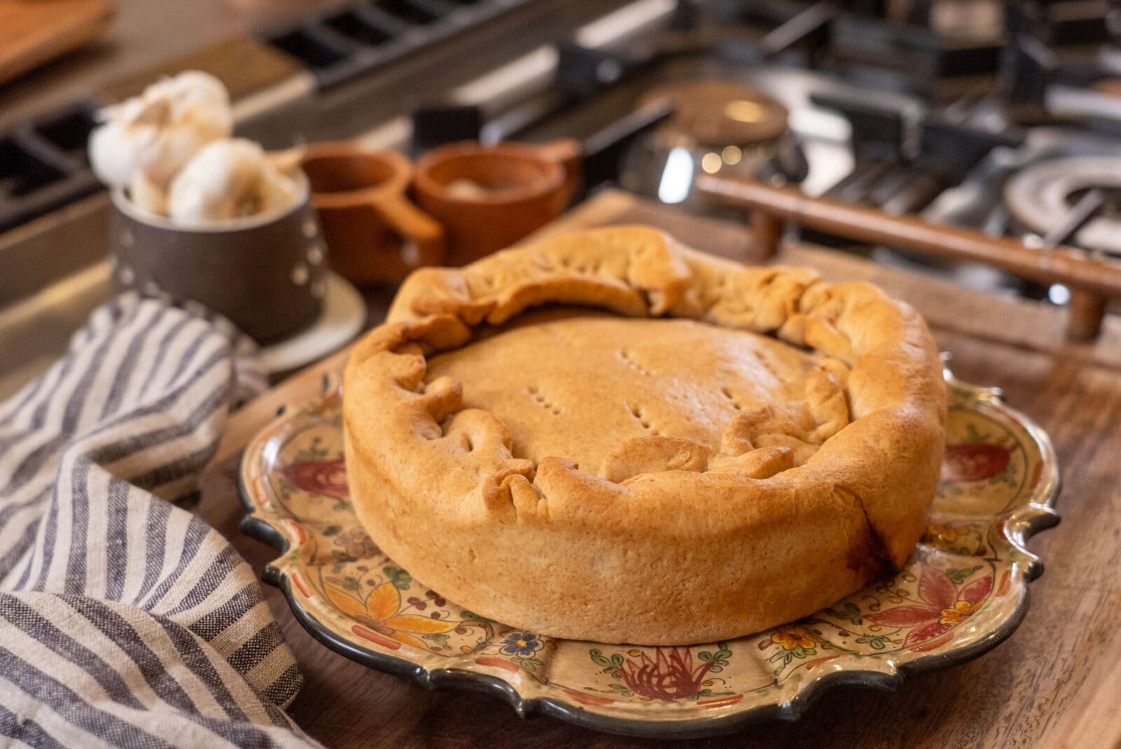 A round, golden-brown pie sits on a decorative plate beside a striped kitchen towel on a wooden surface near a stove.