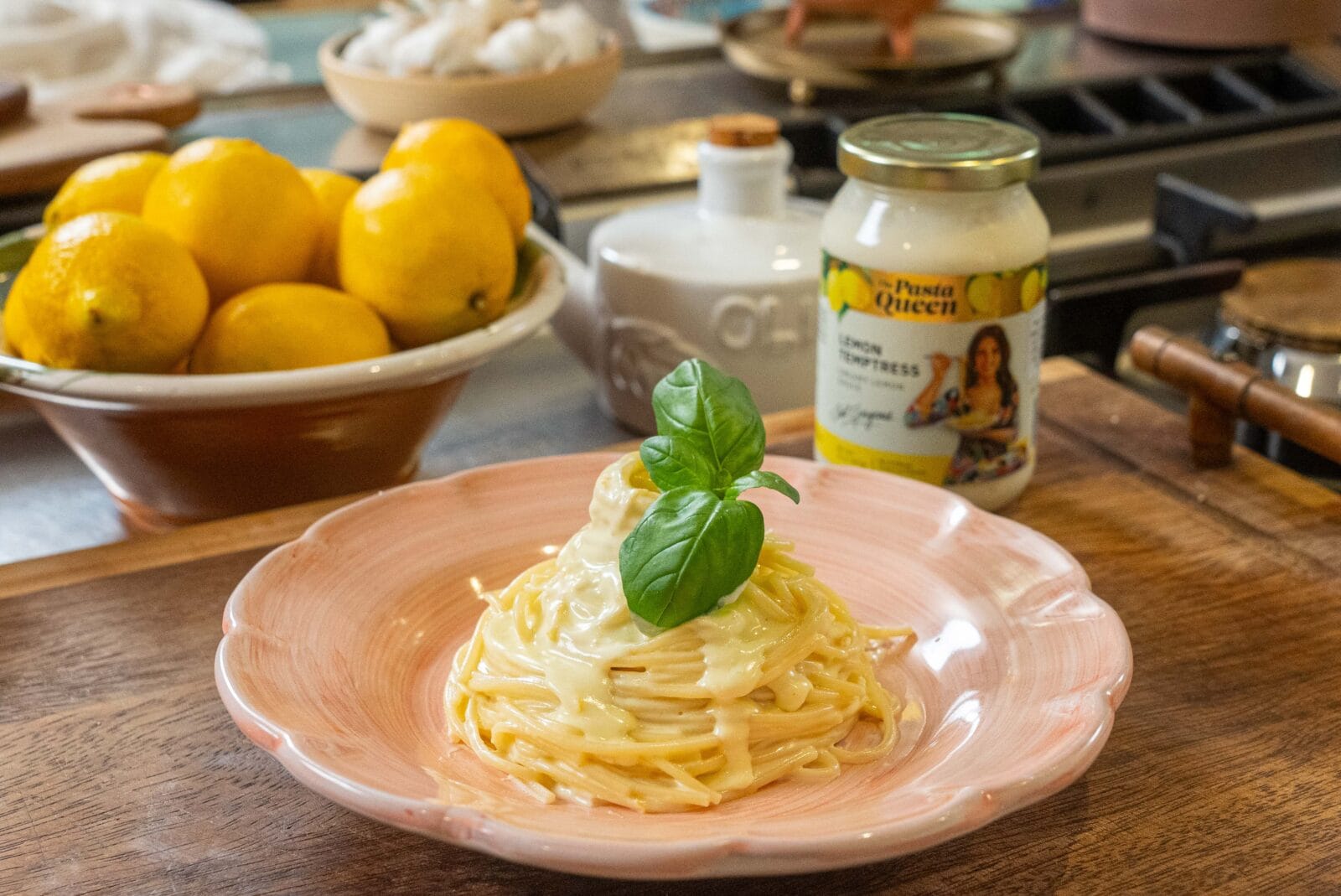 A plate of creamy pasta garnished with basil is set on a wooden counter beside a bowl of lemons and a jar of lemon alfredo sauce.