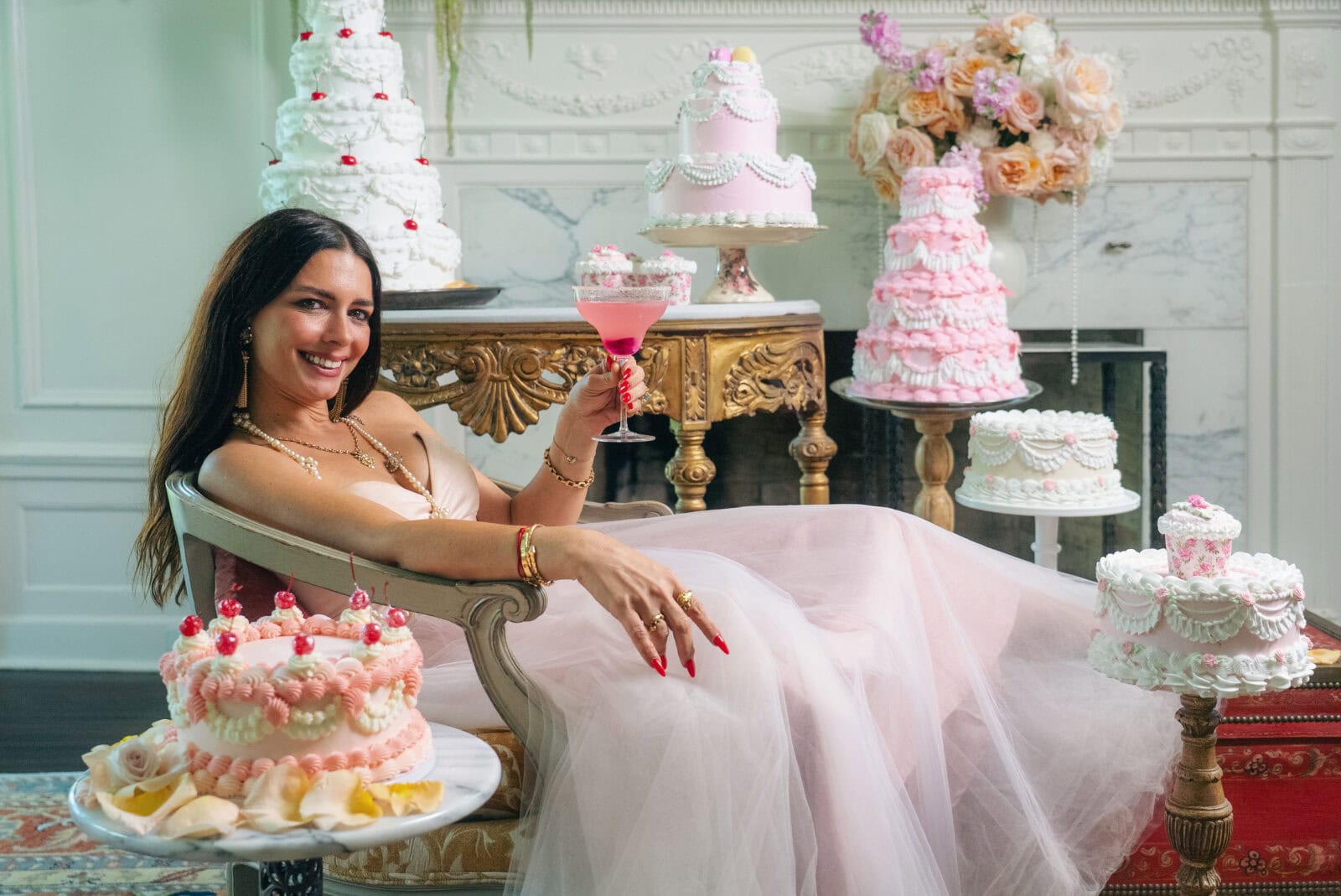 A woman in a pink dress sits on a chaise lounge, holding a pink drink, surrounded by ornate, pastel-colored cakes in an elegant room.