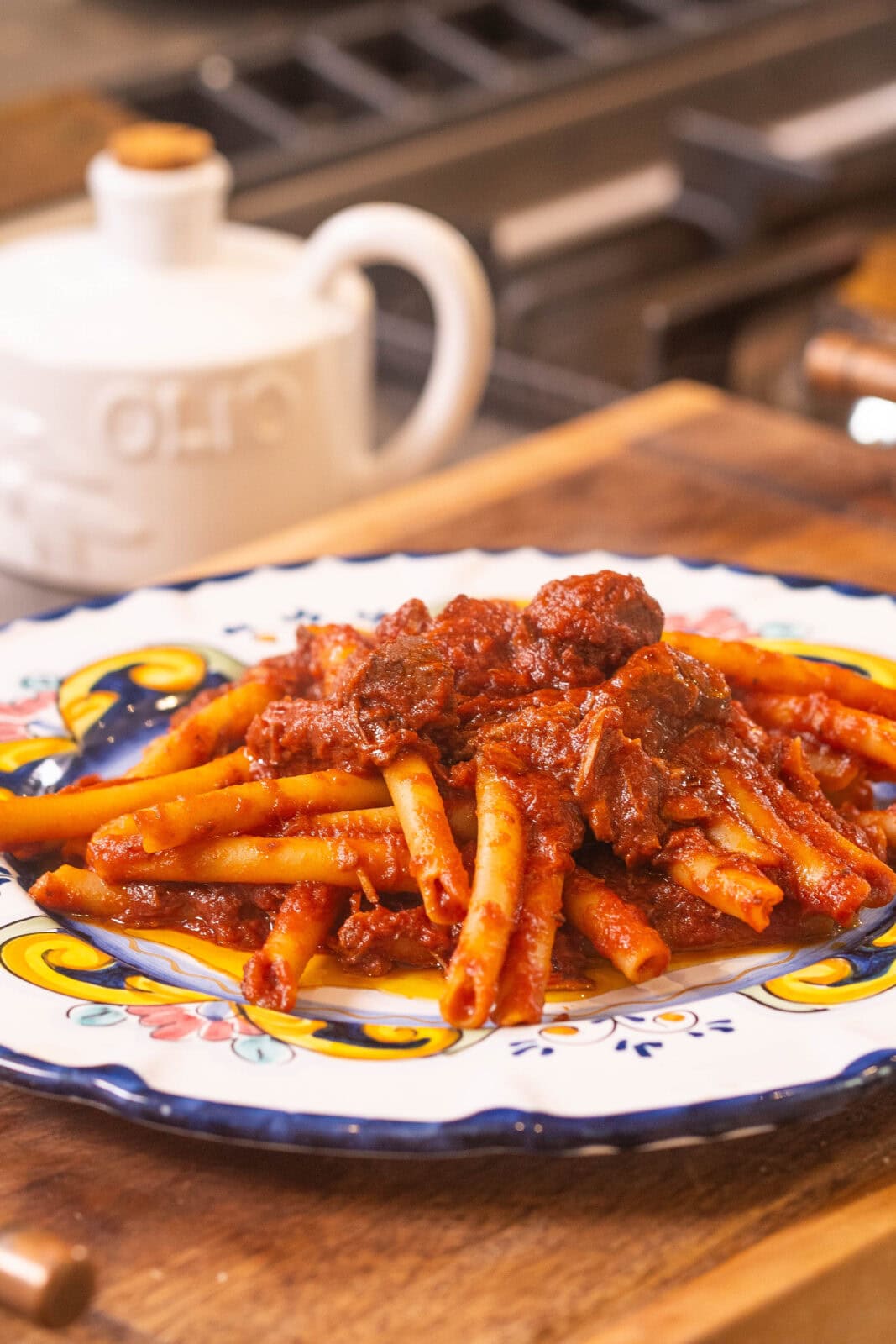A plate of pasta topped with a rich tomato and meat sauce sits on a colorful ceramic dish on a wooden surface, with an olive oil container in the background.