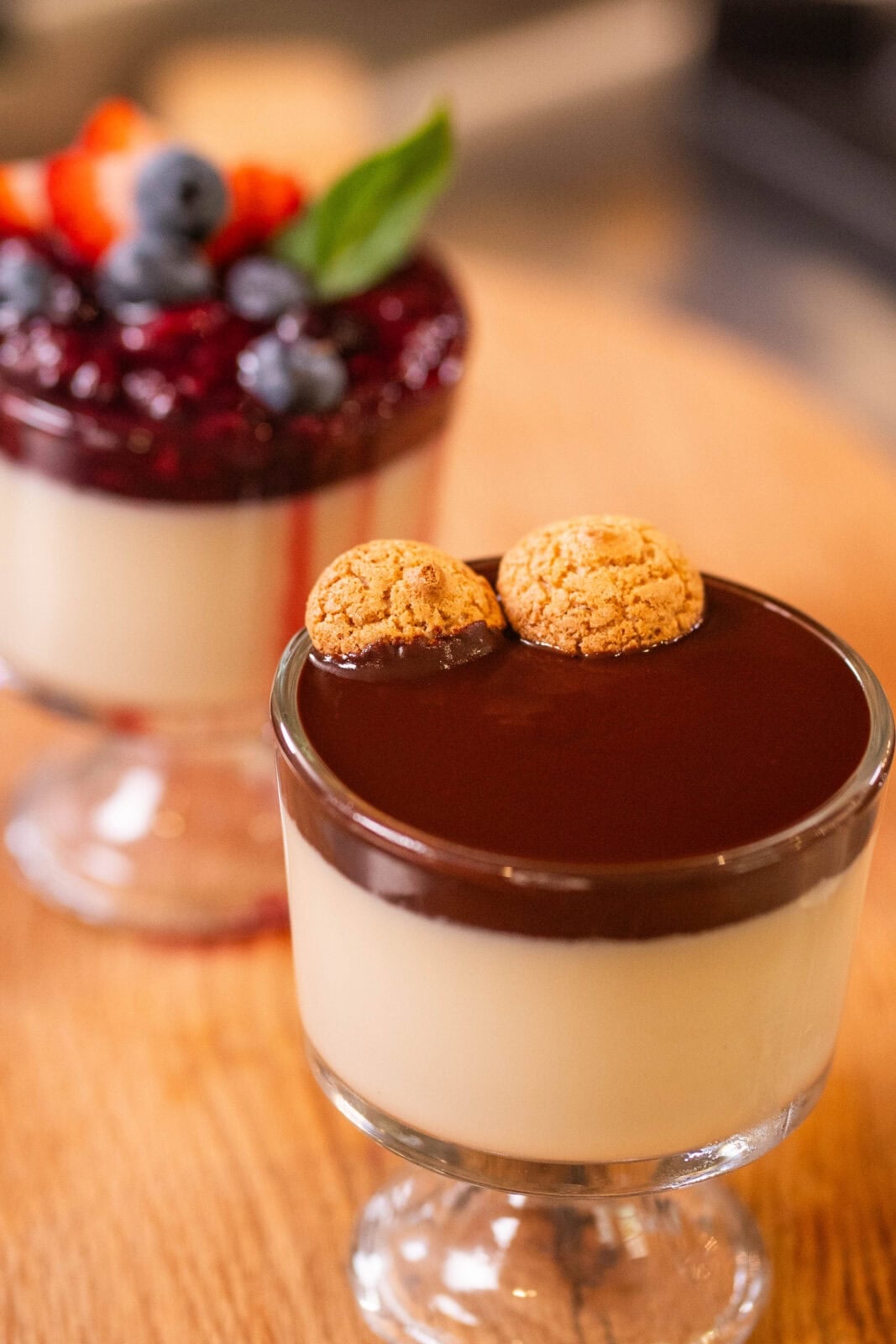Two dessert cups on a wooden surface; one topped with chocolate and biscuits, the other with berry sauce, strawberries, blueberries, and a basil leaf.