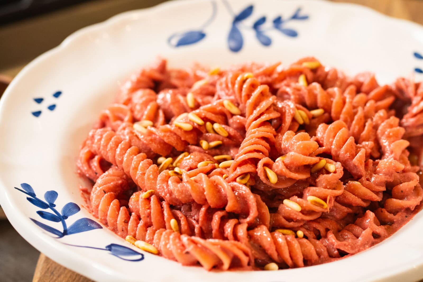 A white plate with blue designs holds fusilli pasta in a pink creamy sauce, garnished with pine nuts.