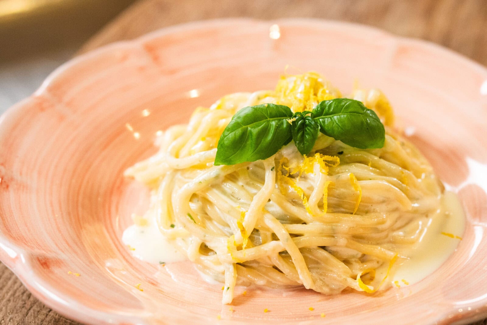 A plate of creamy spaghetti garnished with fresh basil leaves and lemon zest, served on a pink ceramic dish.