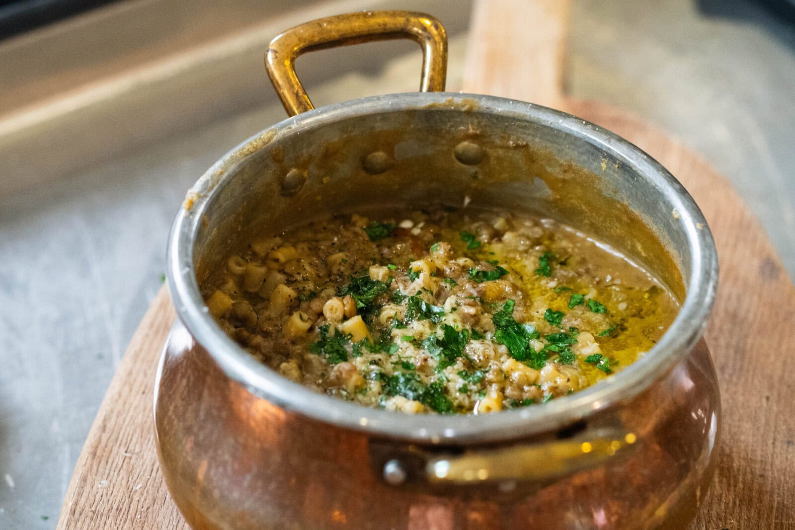 A copper pot filled with a thick, chunky soup or stew containing pasta, herbs, and possibly lentils or minced meat, placed on a wooden surface.