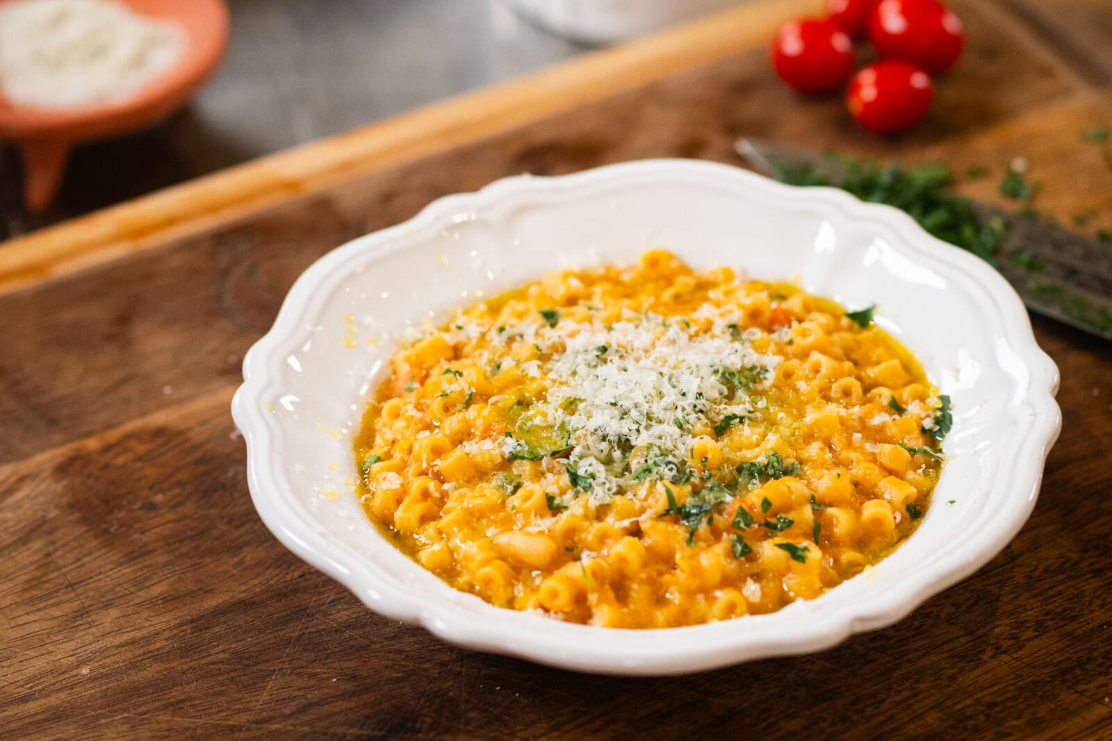 A white bowl filled with pasta in a creamy orange sauce, topped with grated cheese and chopped herbs, placed on a wooden surface.