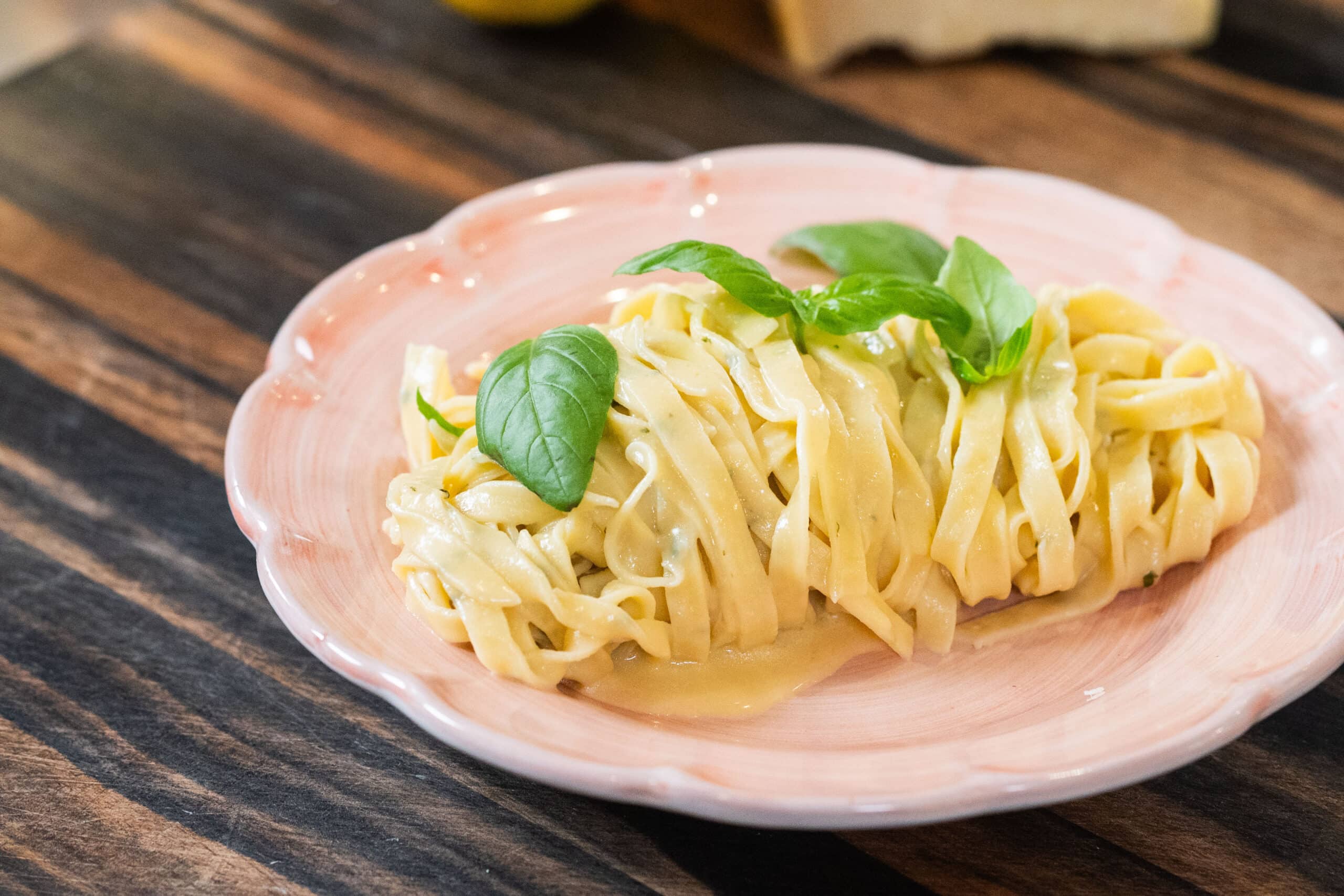 A serving of fettuccine pasta in a creamy sauce, garnished with fresh basil leaves, on a pink plate set on a wooden table.