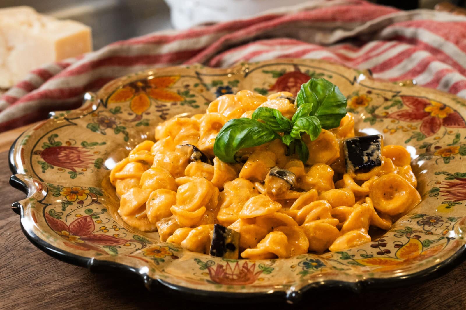 A decorative plate filled with orecchiette pasta in a creamy orange sauce, topped with basil leaves and small pieces of eggplant. A striped cloth and cheese block are in the background.