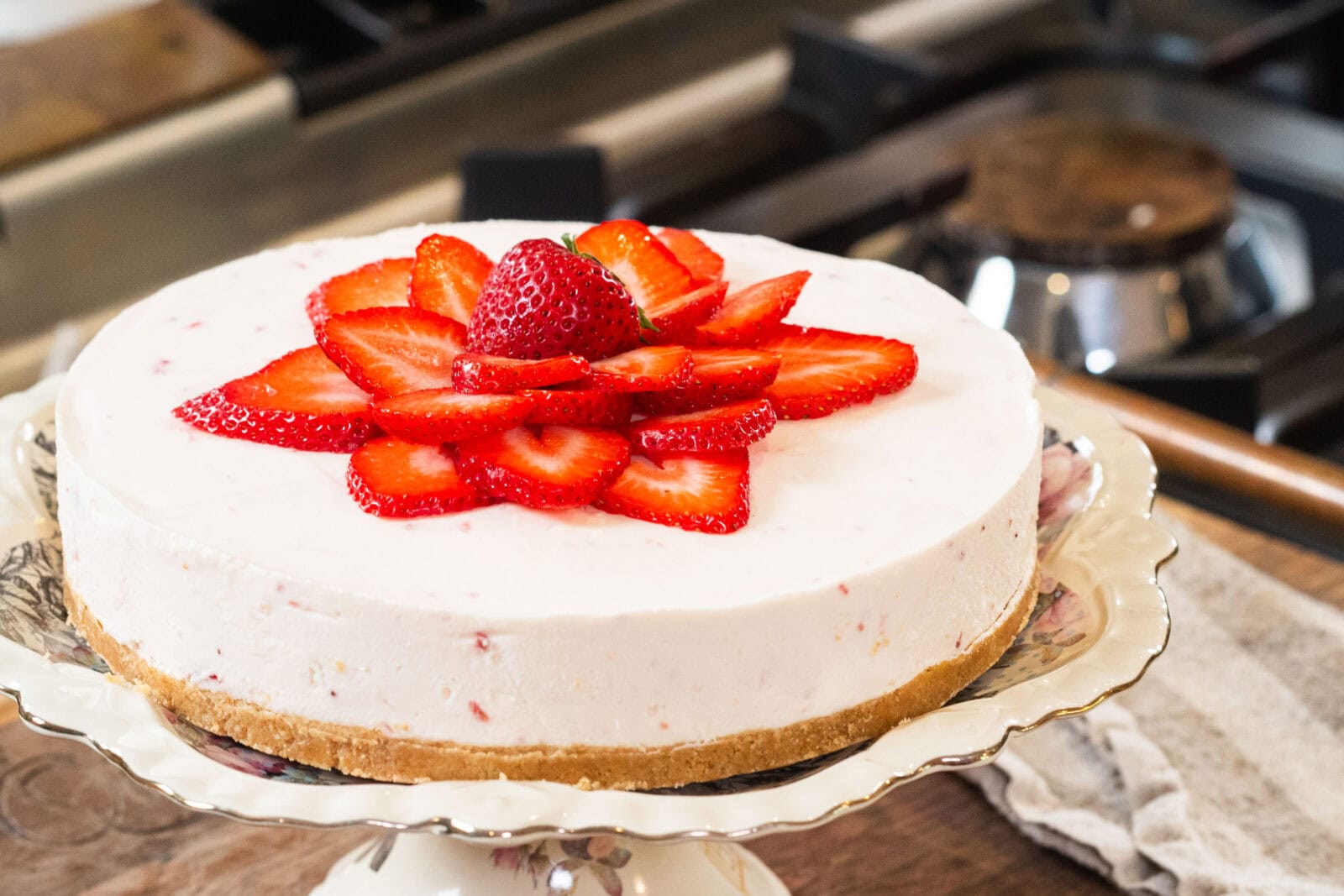 A cheesecake with a graham cracker crust, topped with sliced strawberries and a whole strawberry in the center, displayed on a floral cake stand in a kitchen setting.
