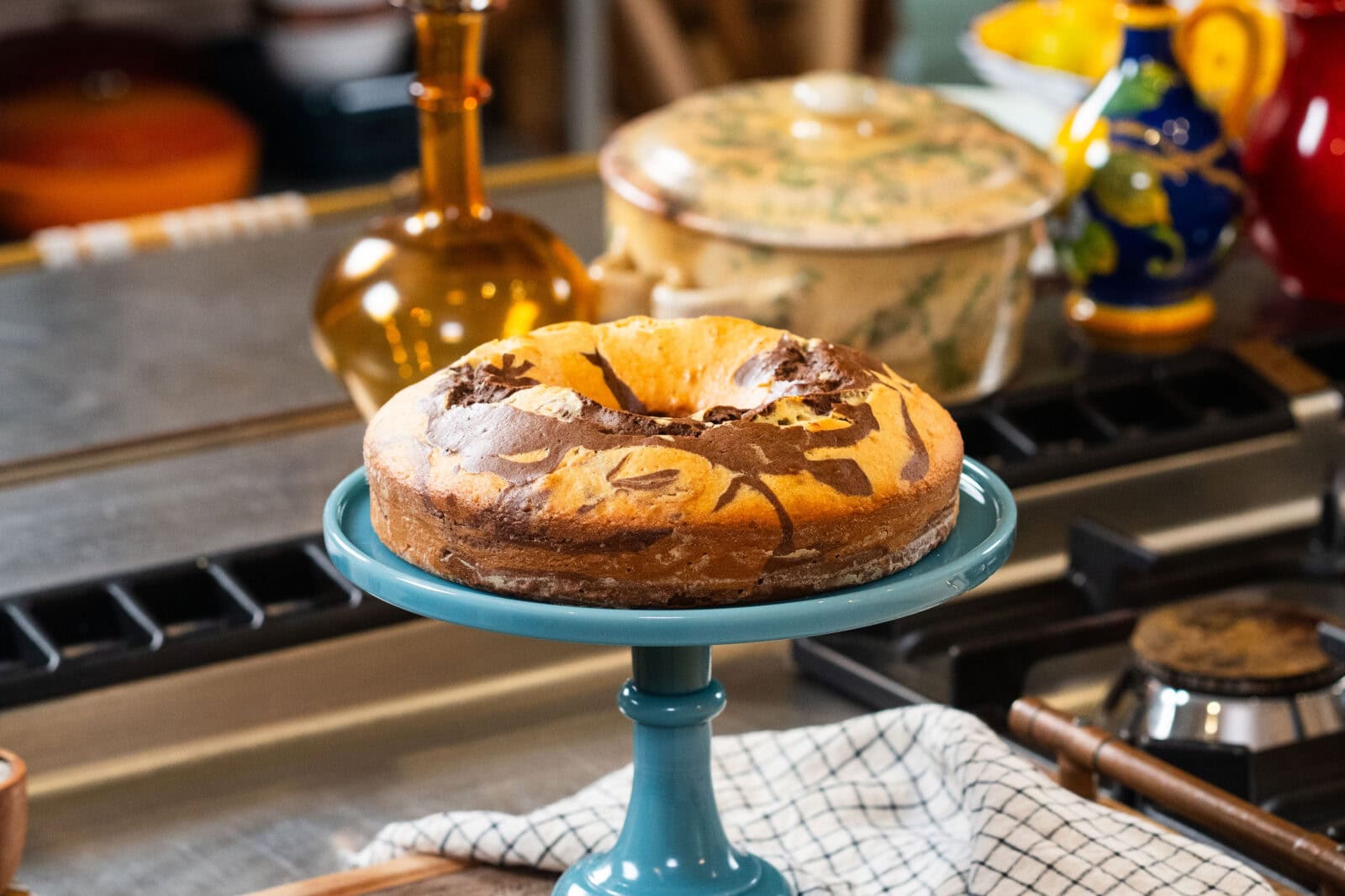 A marble cake with swirled chocolate and vanilla patterns sits on a blue cake stand in a kitchen setting.