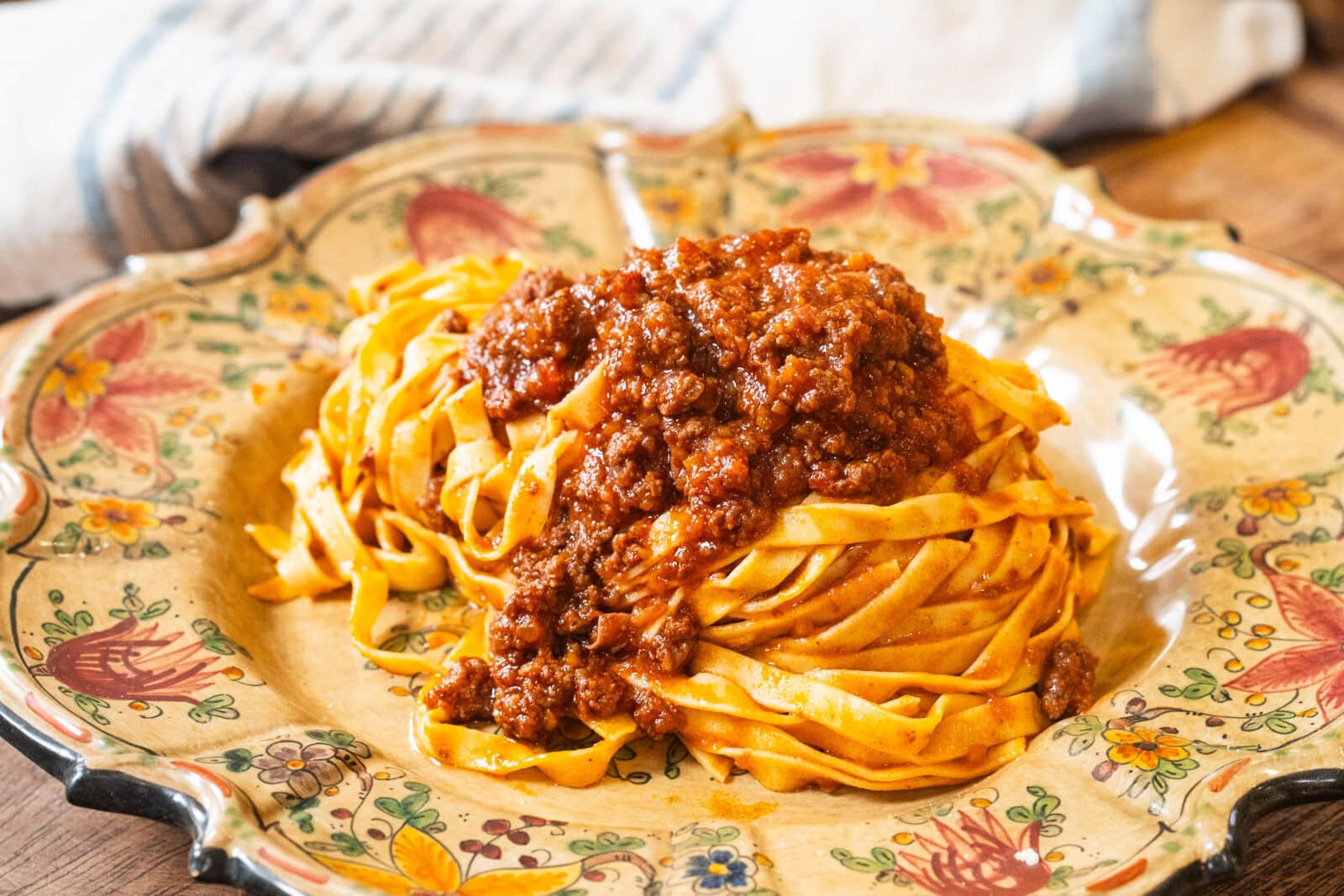 A plate of tagliatelle pasta topped with meat ragù sauce, served on a decorative floral dish with a cloth in the background.
