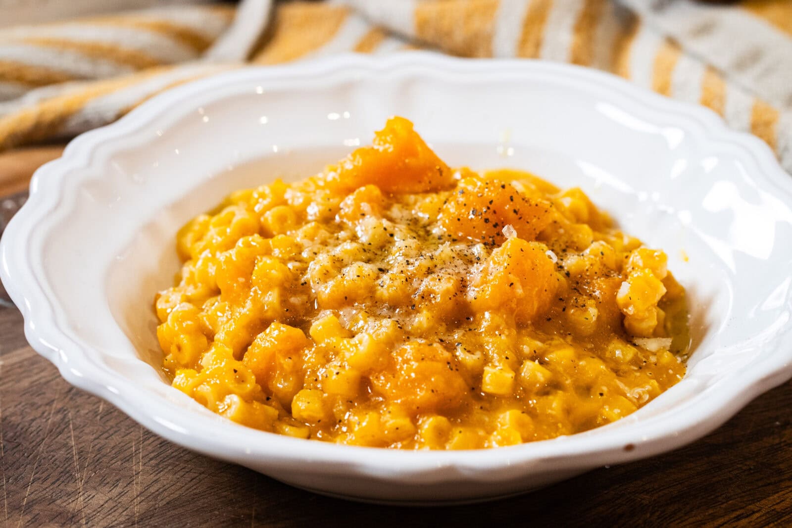 A white bowl filled with creamy pumpkin risotto, garnished with black pepper, sits on a wooden surface with a striped cloth in the background.
