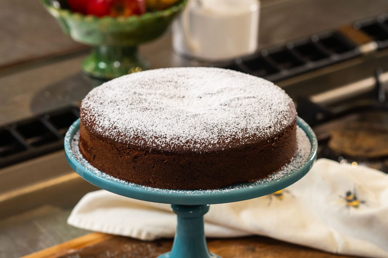 A round chocolate cake dusted with powdered sugar sits on a blue cake stand, with a white cloth and fruit bowl in the background.