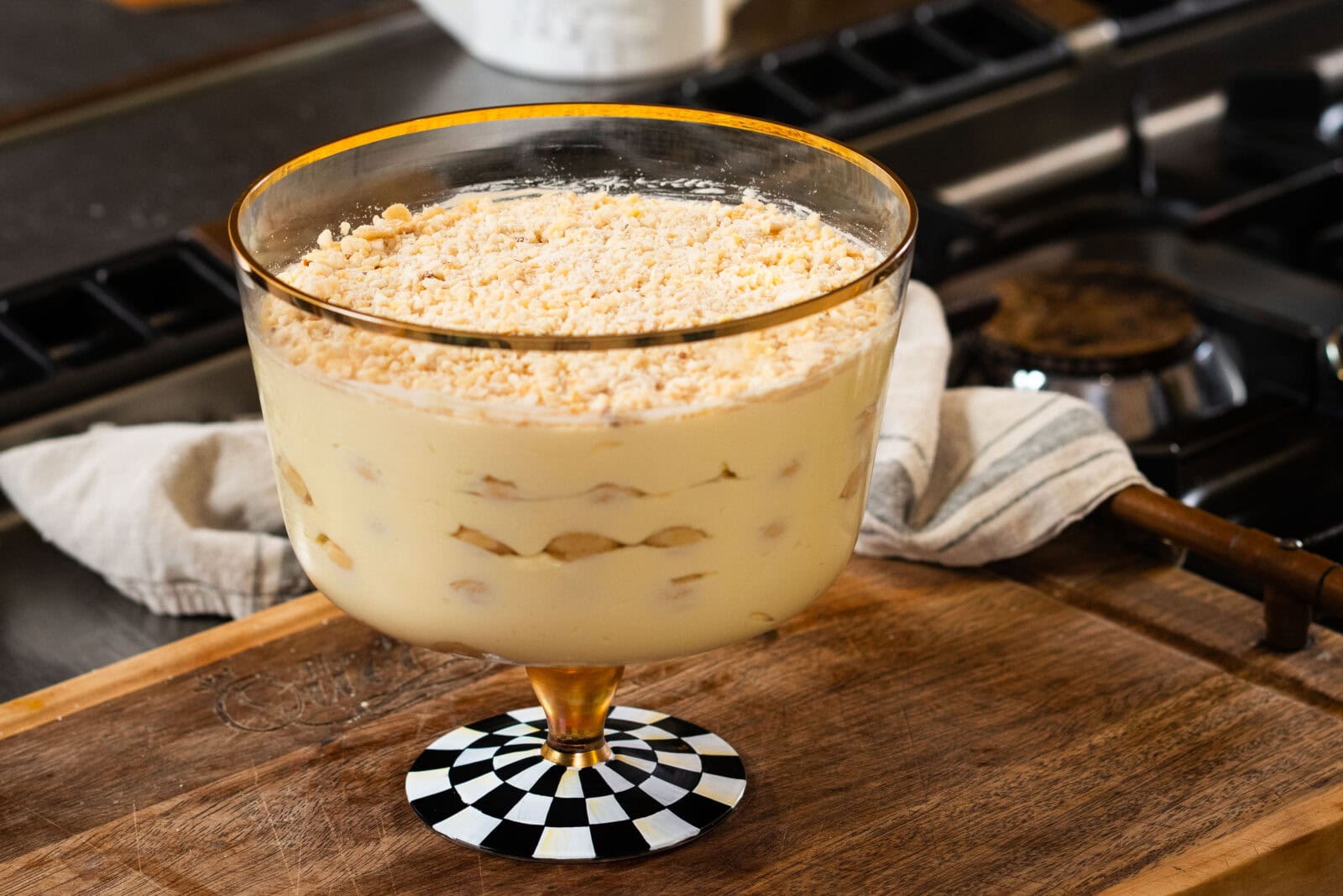 A glass trifle dish filled with layers of creamy pudding, bananas, and crumbled cookies sits on a wooden countertop in a kitchen.
