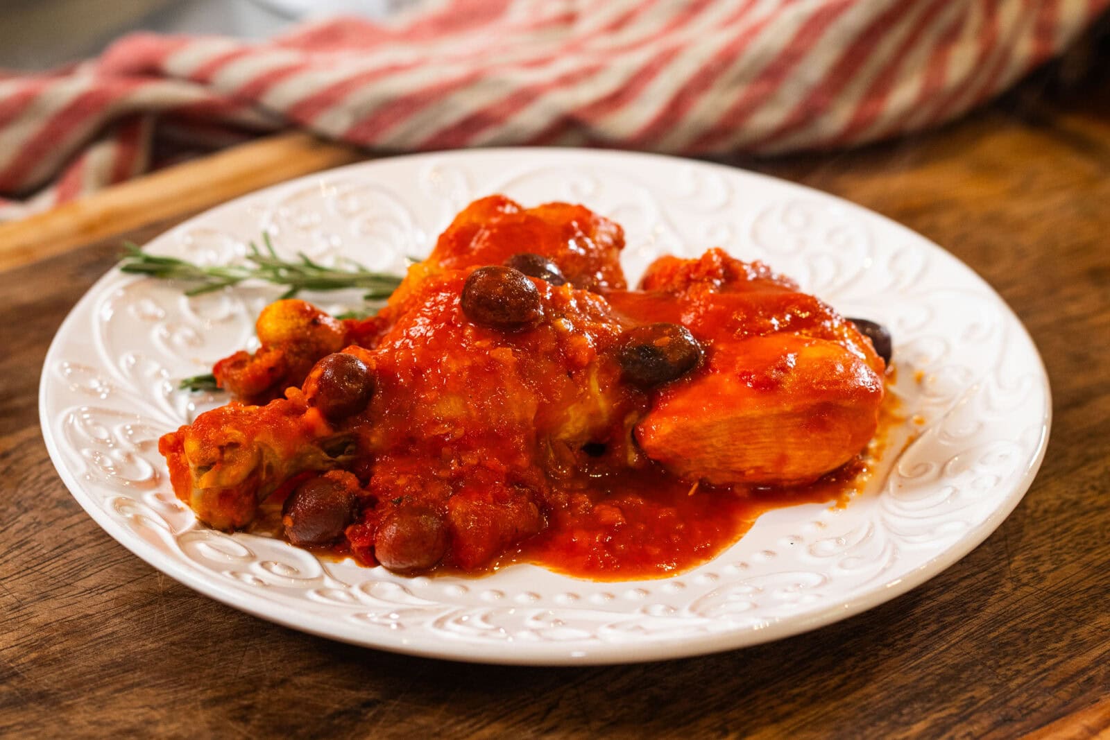 A white plate with chicken in red tomato sauce, garnished with olives and herbs, sits on a wooden surface with a striped cloth in the background.