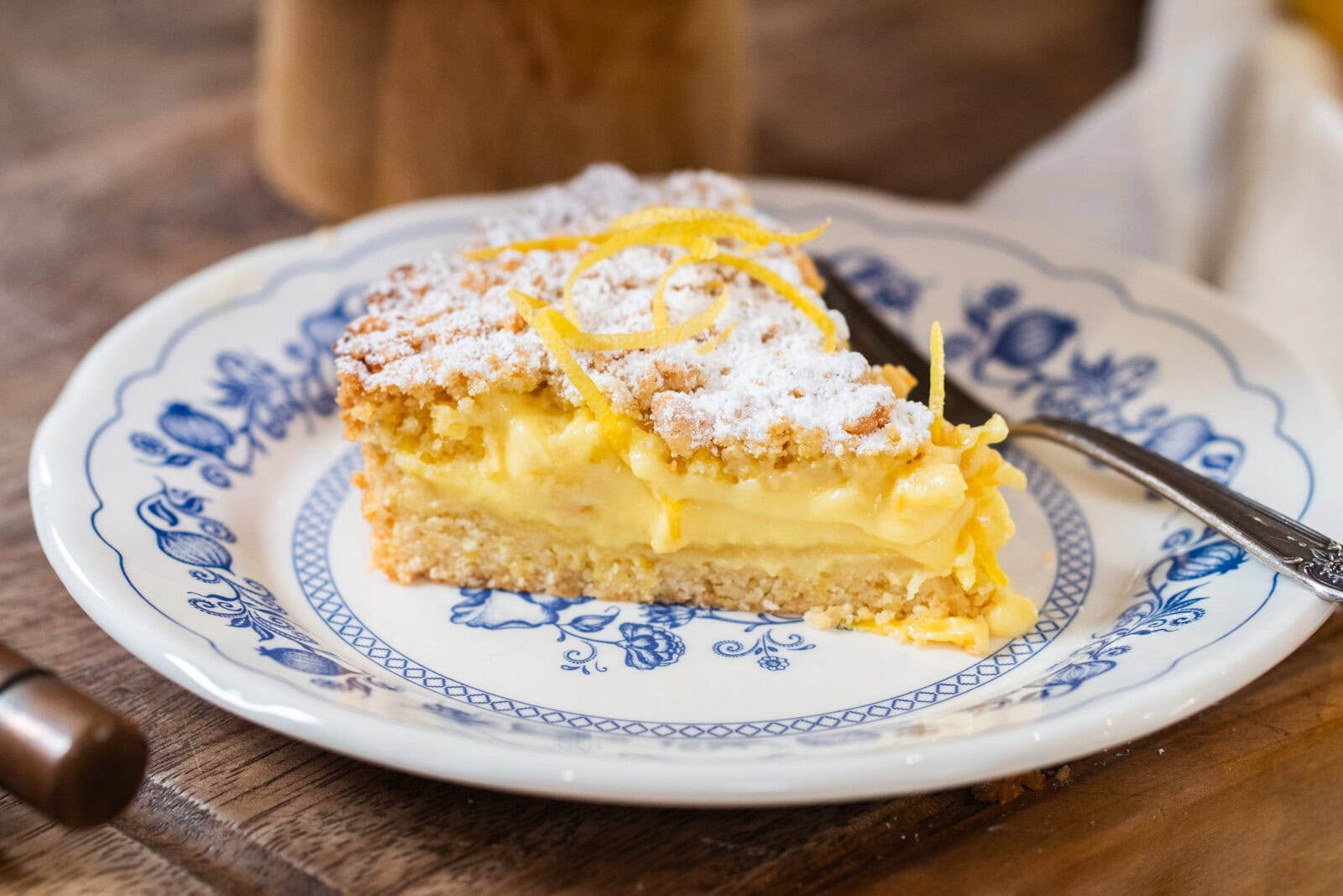 A slice of lemon cream pie with powdered sugar and lemon zest on top, served on a blue and white plate with a fork.
