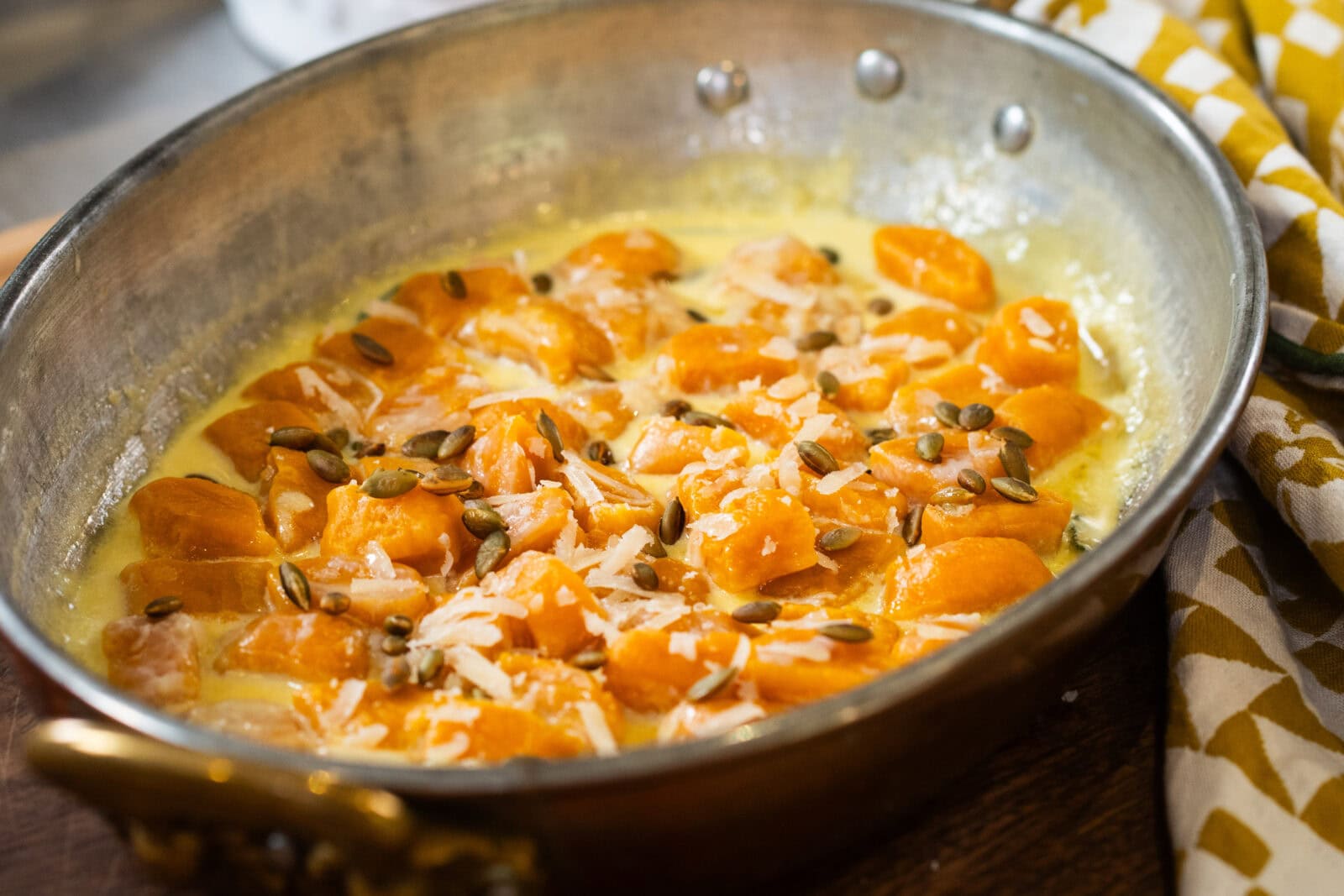 A metal pan contains cooked gnocchi in a creamy sauce, topped with grated cheese and pumpkin seeds.