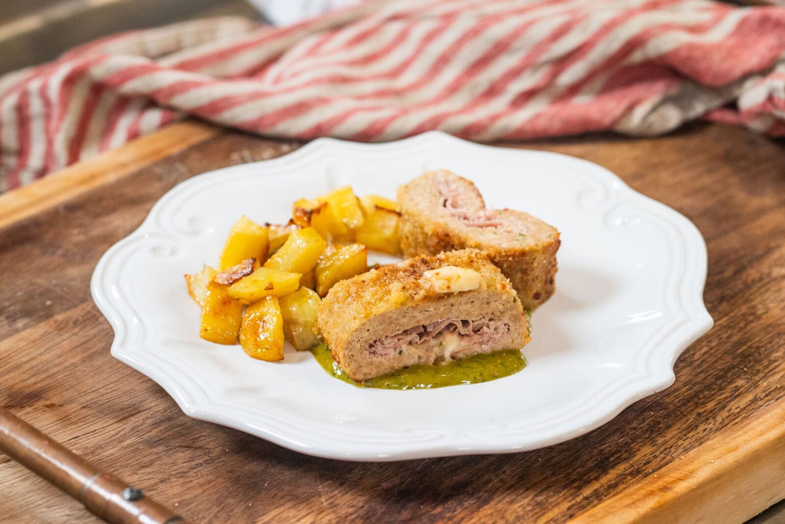 A white plate with breaded ham and cheese rolls sliced in half, served with roasted potatoes and a green sauce on a wooden surface.