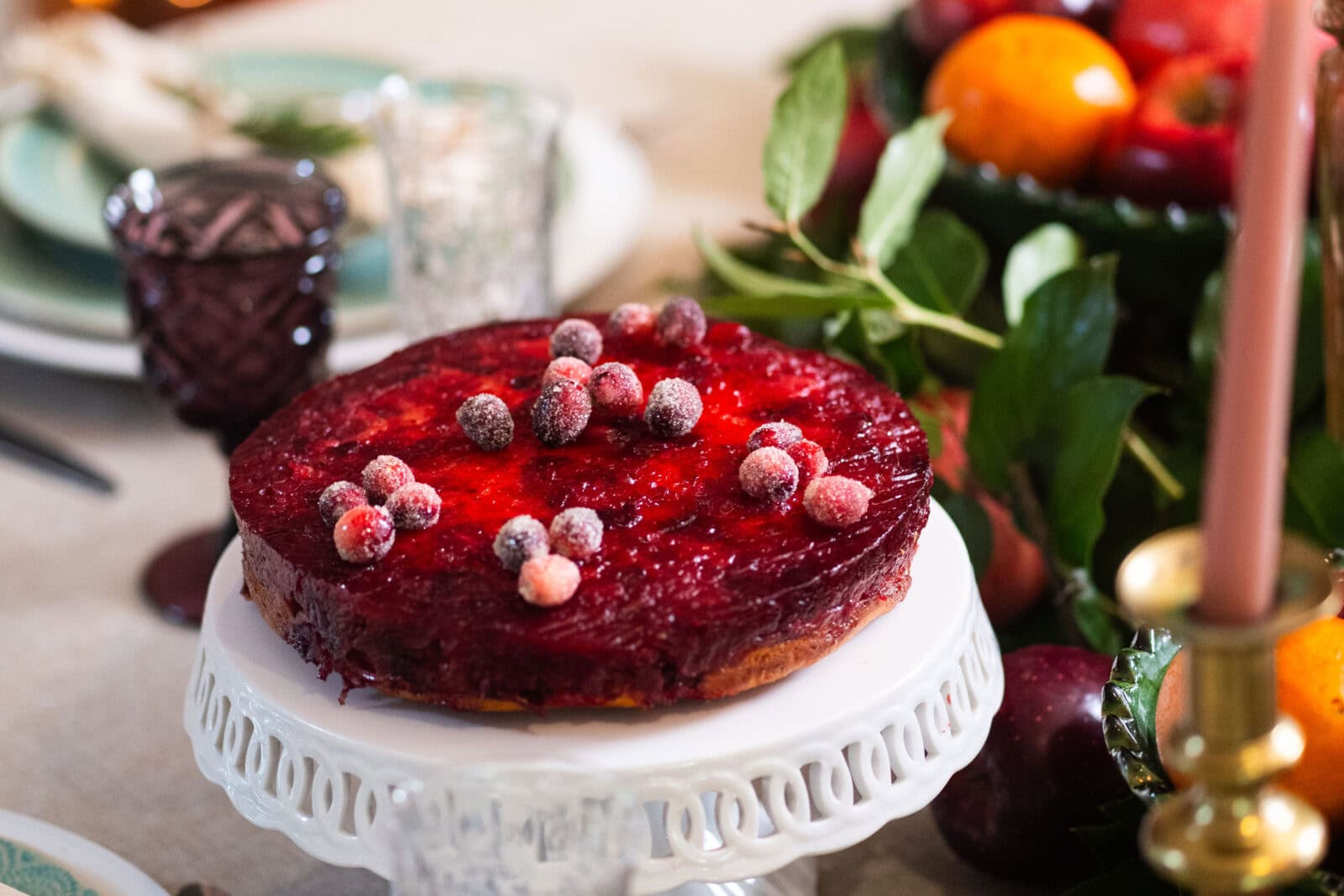 A round cranberry-topped cake on a white stand sits on a table set for a meal, with fruit and greenery in the background.