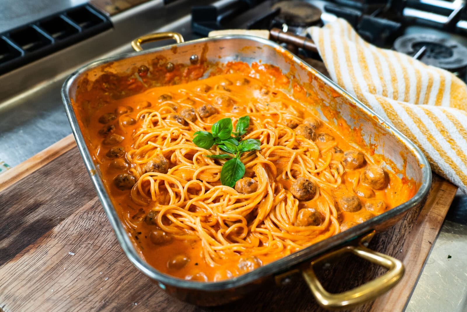 A baking dish filled with spaghetti and meatballs in a creamy tomato sauce, garnished with fresh basil, sits on a wooden board near a stove.