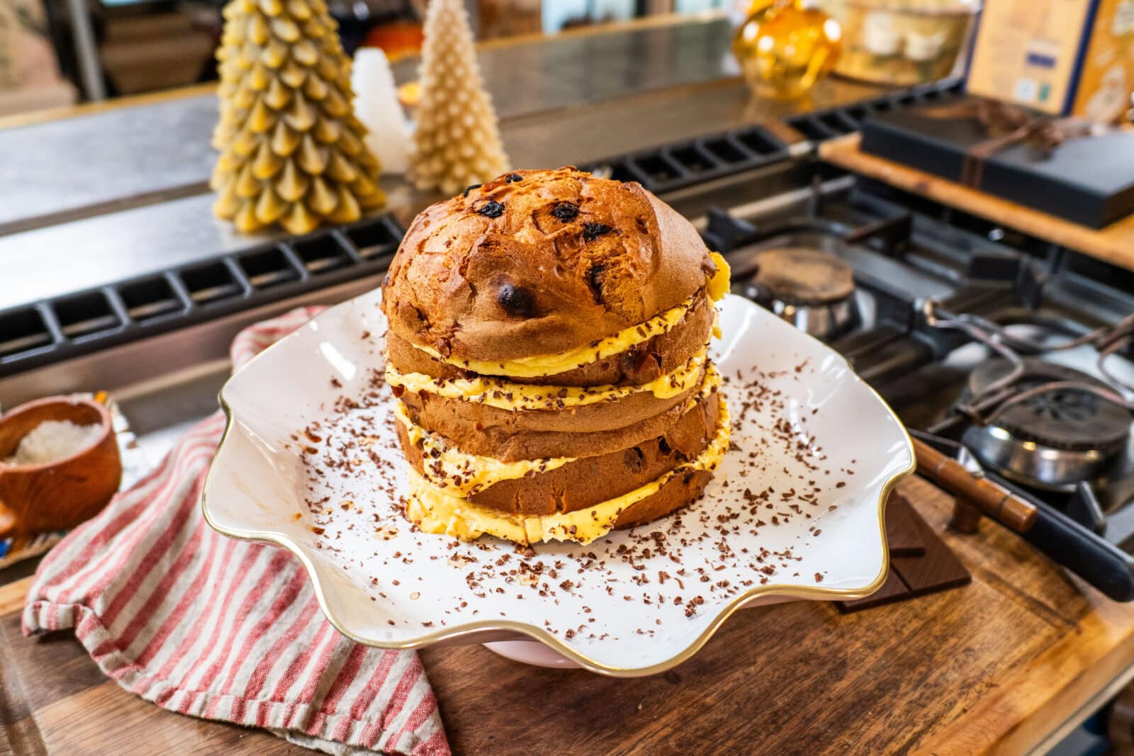 A sliced panettone cake with cream filling and chocolate shavings sits on a decorative plate on a kitchen counter.