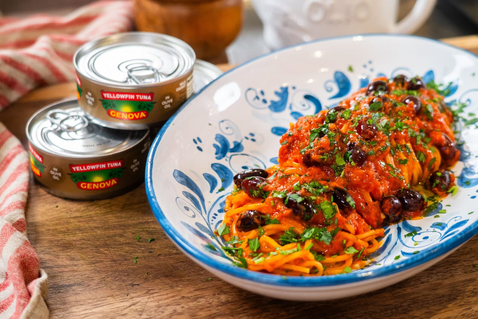 A plate of spaghetti topped with tomato sauce, black olives, and herbs sits beside two cans of Genova yellowfin tuna on a wooden surface.