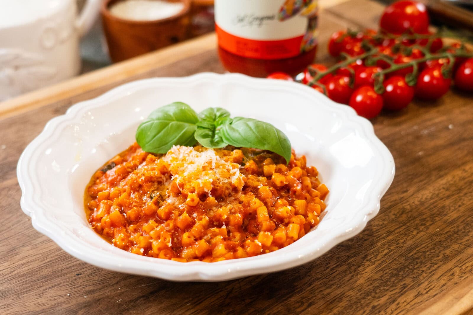 A white bowl of tomato-based pasta garnished with basil and grated cheese sits on a wooden table, with cherry tomatoes and a sauce bottle in the background.