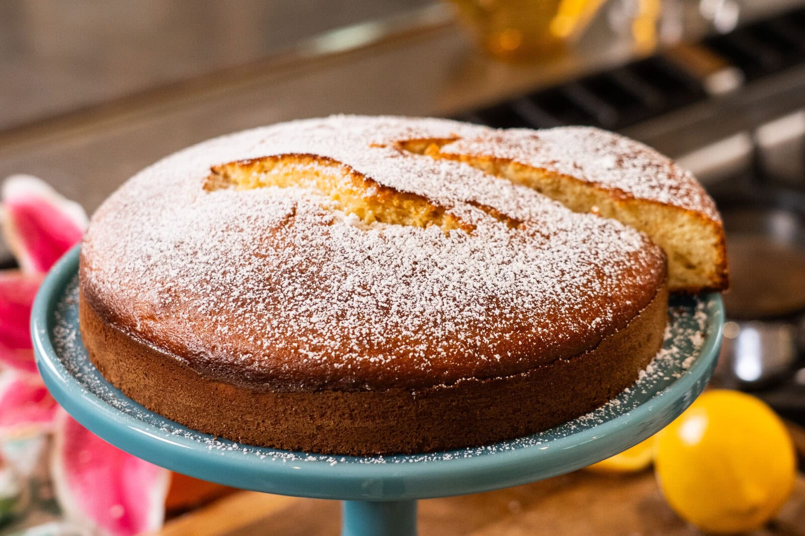 A round, golden-brown cake dusted with powdered sugar sits on a blue cake stand, with one slice cut and slightly separated from the whole cake.