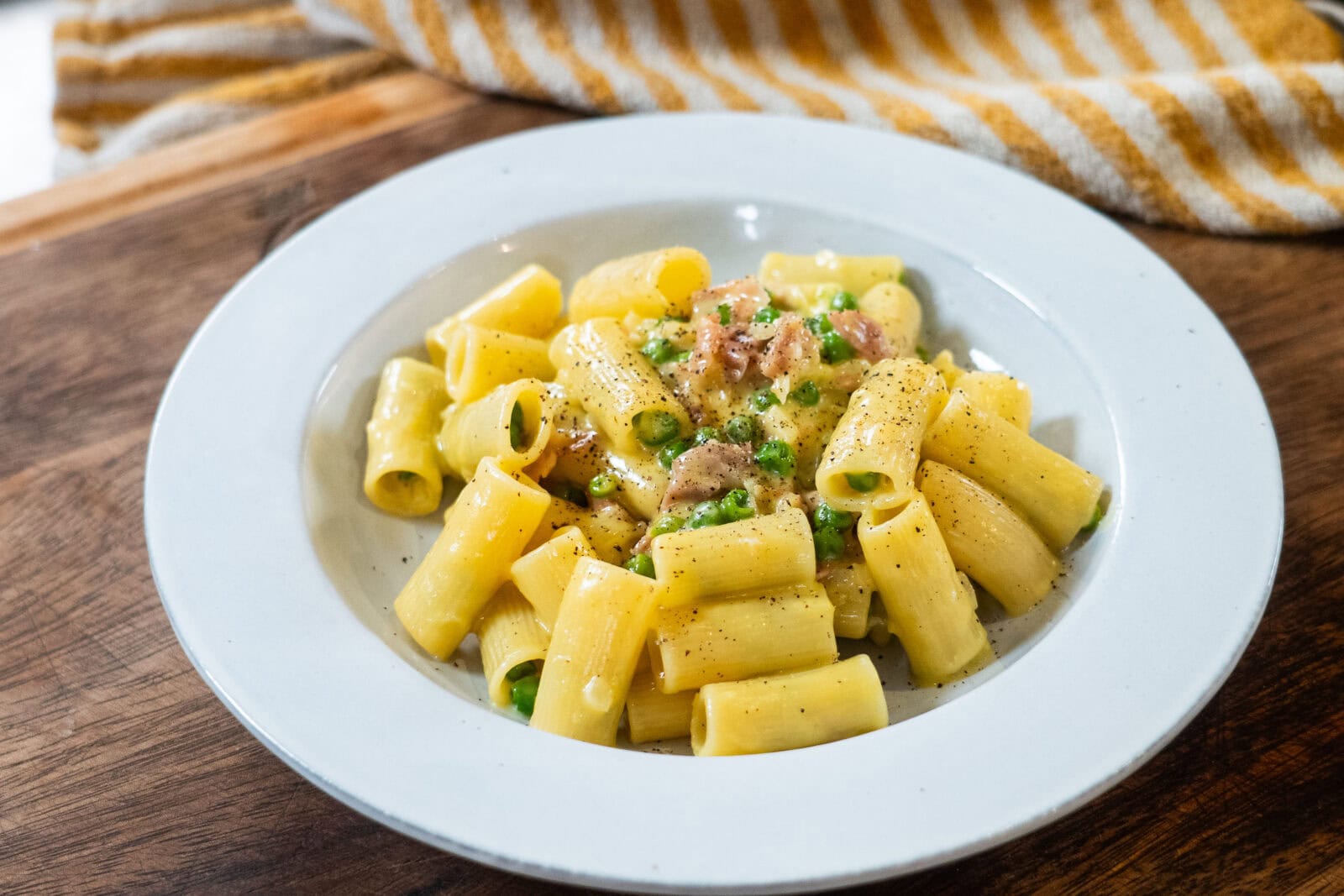 A white plate of rigatoni pasta with peas, ground meat, and a creamy sauce, garnished with black pepper, on a wooden surface.