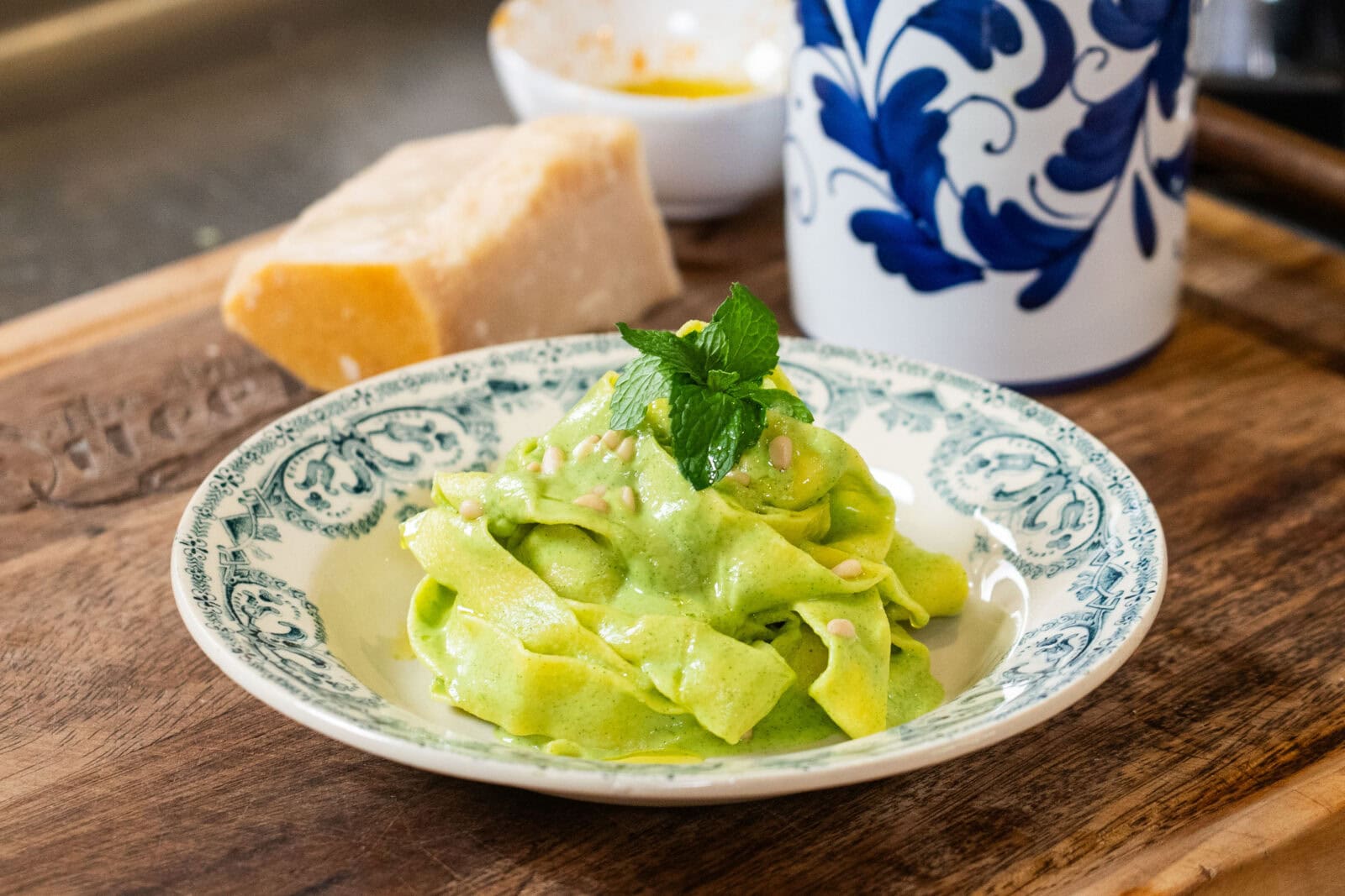 A plate of pasta with green sauce is garnished with mint leaves on a wooden surface, alongside a block of cheese and a blue-and-white container.