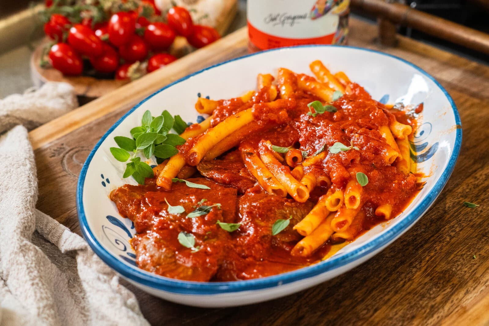 A white oval dish with ziti pasta and meat covered in tomato sauce, garnished with fresh herbs, sits on a wooden surface.