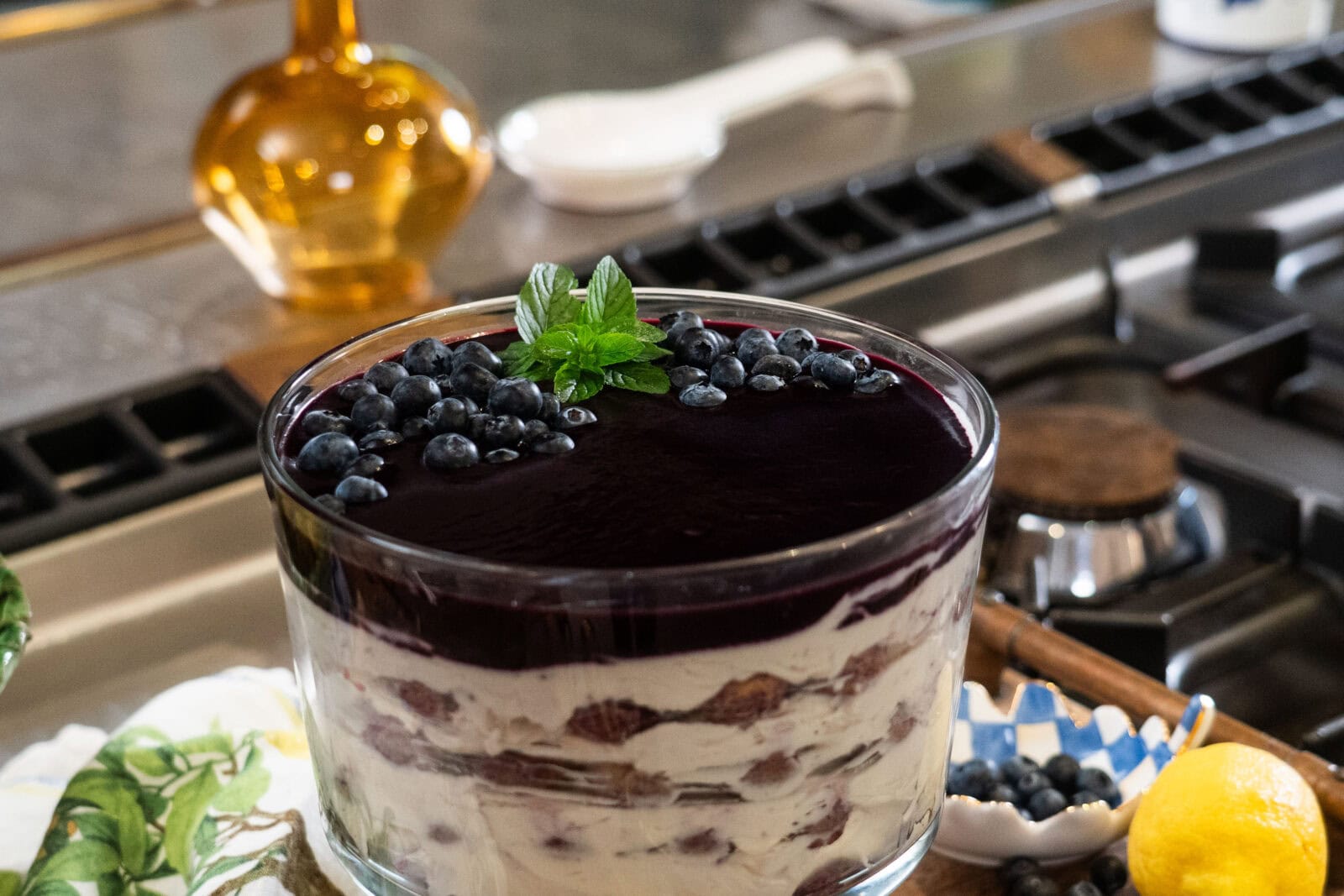 A layered trifle dessert with cream, cake, and blueberry topping garnished with mint leaves, sitting on a kitchen counter near a bowl of fresh blueberries.
