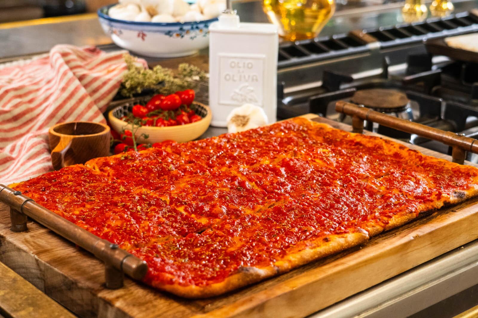 Rectangular sheet pan pizza with tomato sauce sits on a wooden board in a kitchen, surrounded by ingredients like garlic, tomatoes, and olive oil.