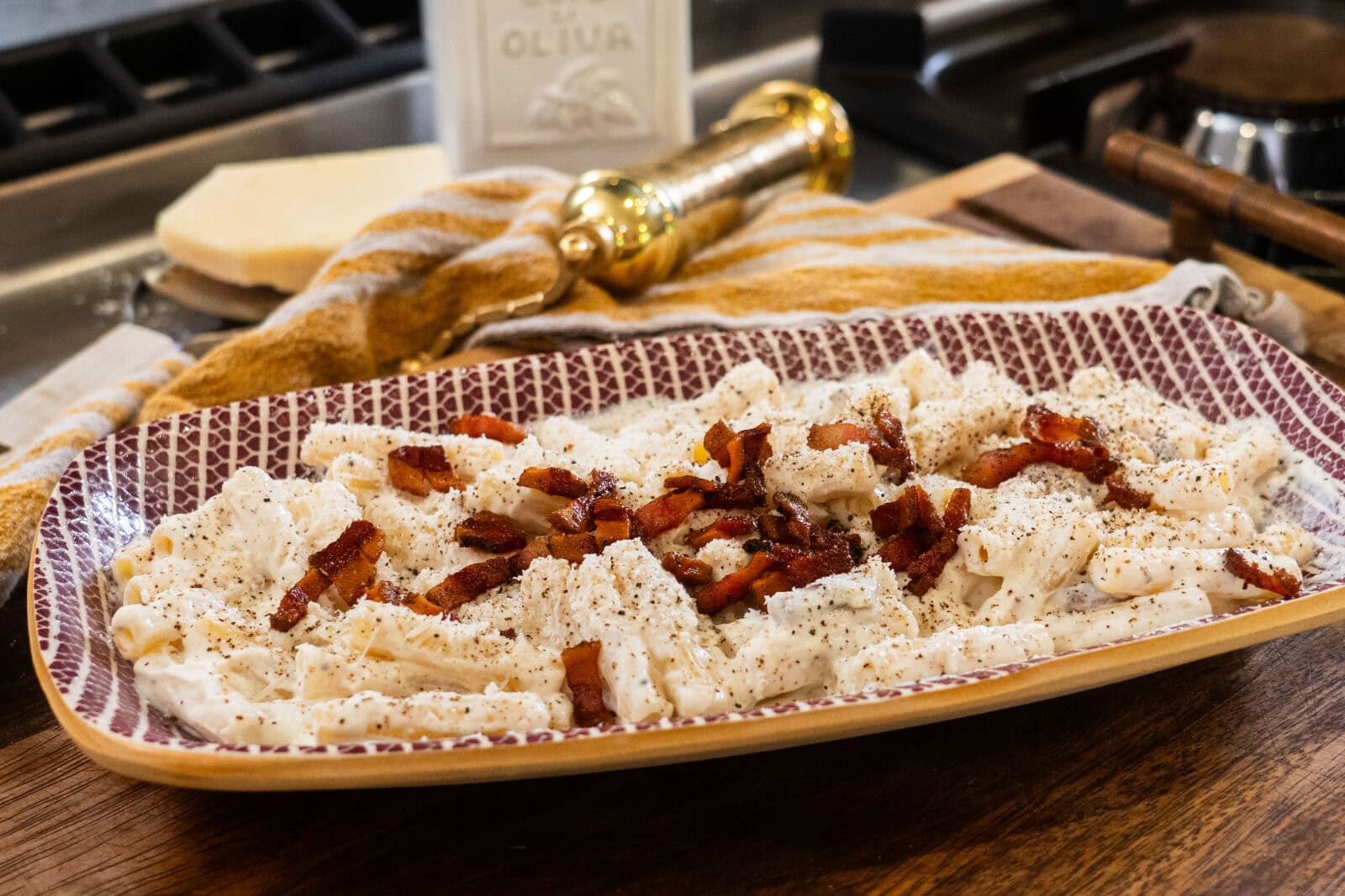 A rectangular plate of creamy pasta topped with crumbled bacon and black pepper is on a wooden surface, with a cheese wedge, a gold grinder, and a striped towel in the background.