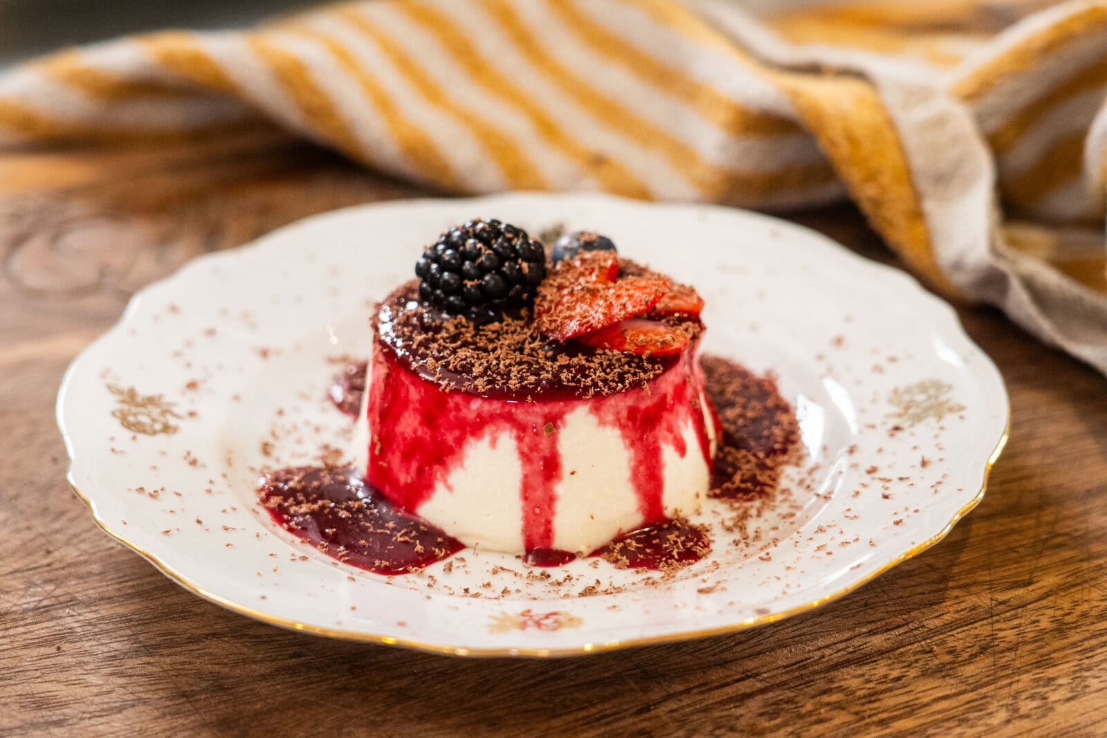 A dessert of panna cotta with berry sauce, topped with sliced strawberries, a blackberry, and chocolate shavings, served on a decorative white plate.