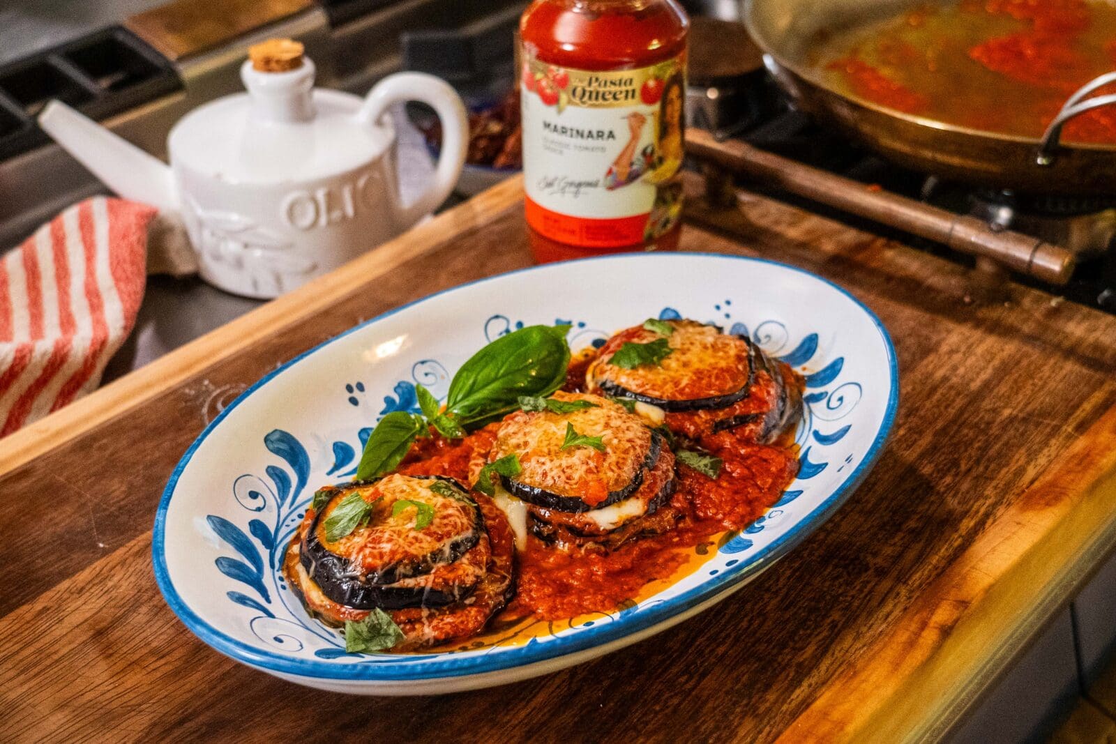 Three baked eggplant parmesan stacks with marinara sauce and melted cheese, garnished with basil, served on a decorative plate next to a jar of marinara sauce and a cooking pan.