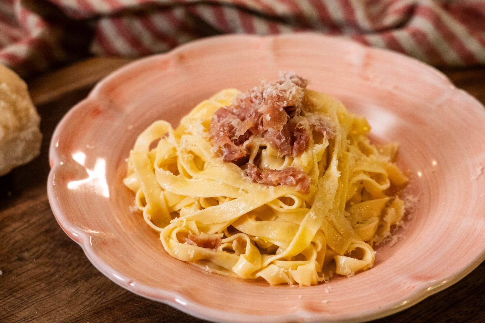 A plate of tagliatelle pasta topped with grated cheese and pieces of ham sits on a pink dish with a striped cloth in the background.