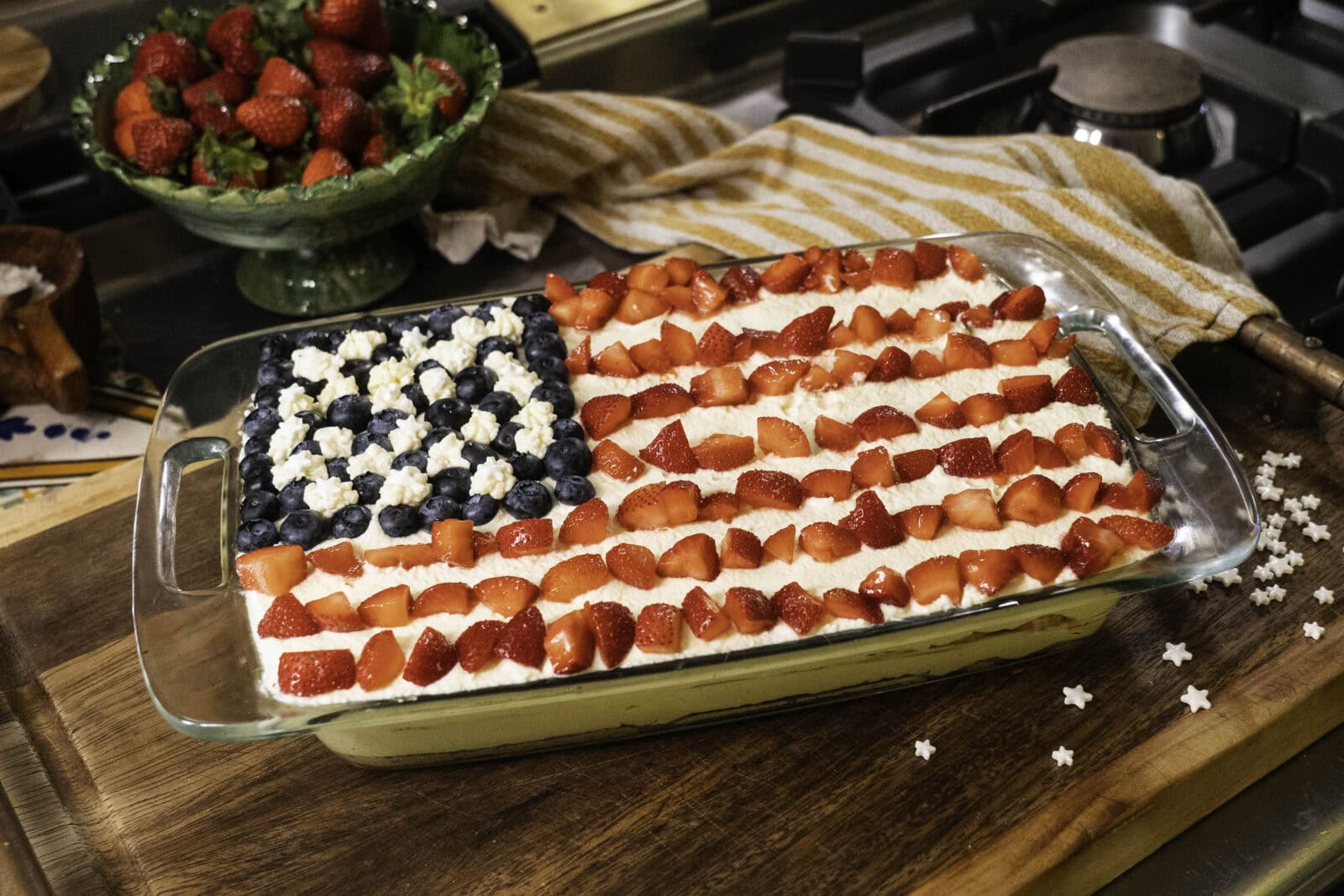 A rectangular dessert decorated like the American flag with blueberries, strawberries, and white cream sits on a wooden surface near a bowl of strawberries and a striped towel.