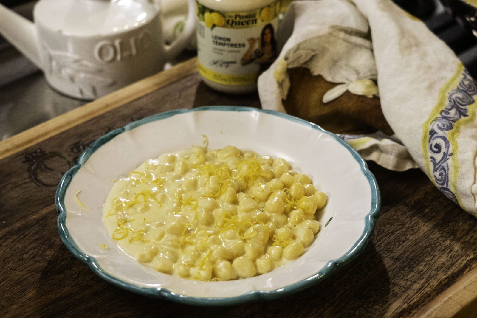 A white bowl with green trim contains creamy pasta shells topped with lemon zest, set on a wooden surface beside a loaf of bread and a jar labeled "Lemon Empress.
